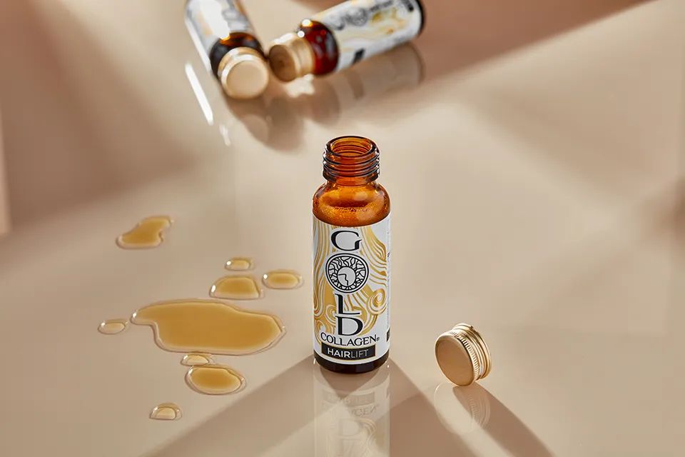 Braune Glasflasche mit Gold Collagen Hairlift-Etikett. Offener Deckel daneben. Flüssigkeit daneben verschüttet.