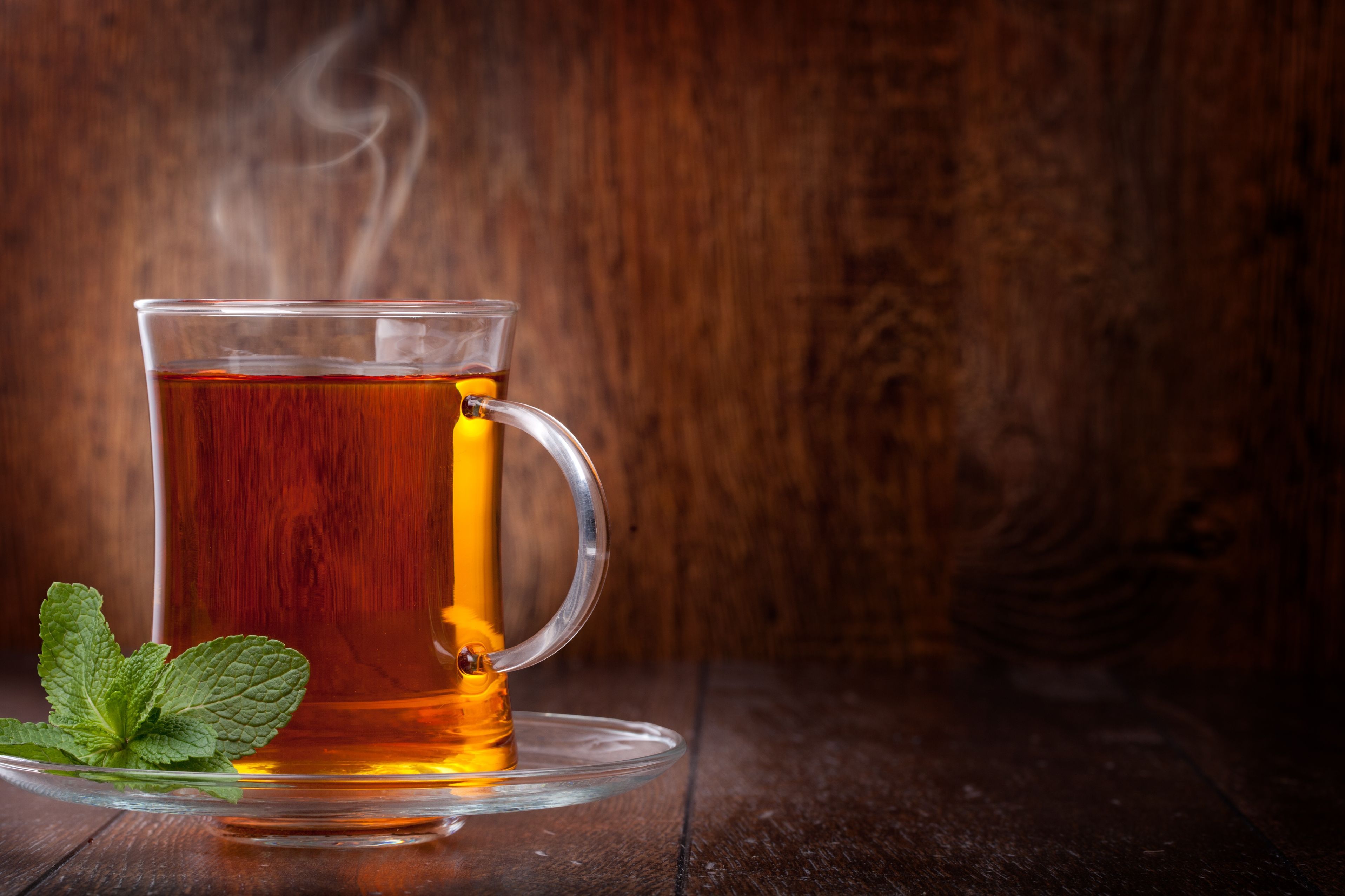 Tasse Tee mit Minze. Glasbecher mit dampfendem Tee auf Untertasse, daneben Minzblätter. Holz Hintergrund.
