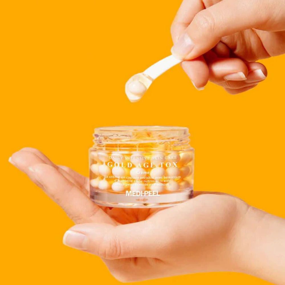 Glasbehälter mit gelben Kügelchen, gehalten von Händen. Löffel entnimmt eine Kugel. Aufschrift: Gold Age Tox, Medipeel.