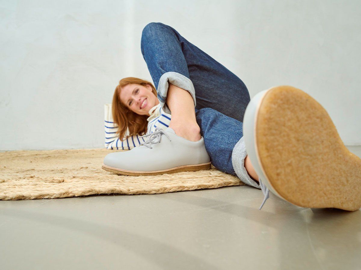 Frau mit hellgrauen Schnürschuhen. Breite Passform. Auf Teppich liegend, Jeans, gestreiftes Oberteil.