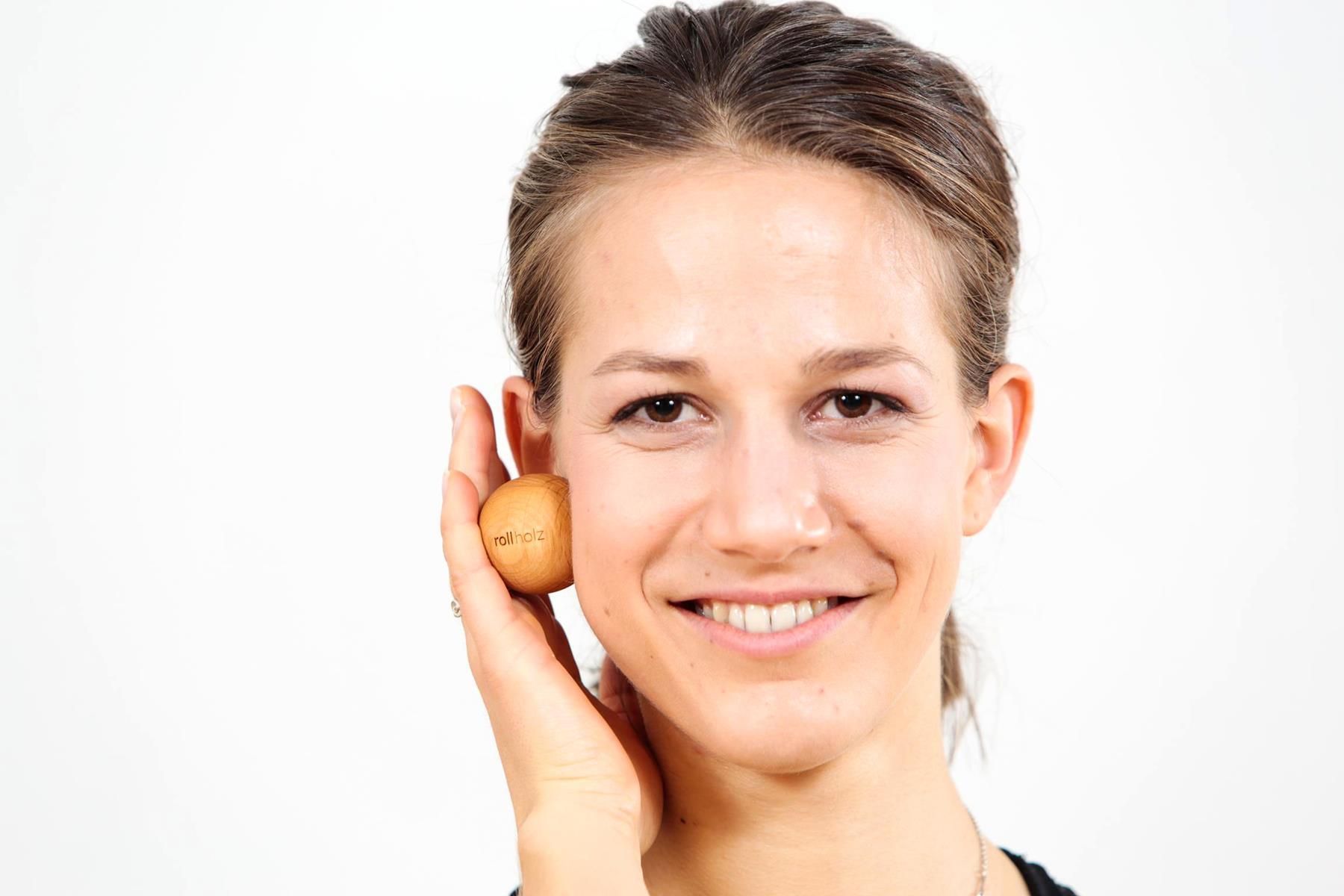 Eine Frau hält einen kleinen Faszienball aus Holz an ihrer Wange. Der Ball ist aus Walnussholz und trägt die Aufschrift rollholz.