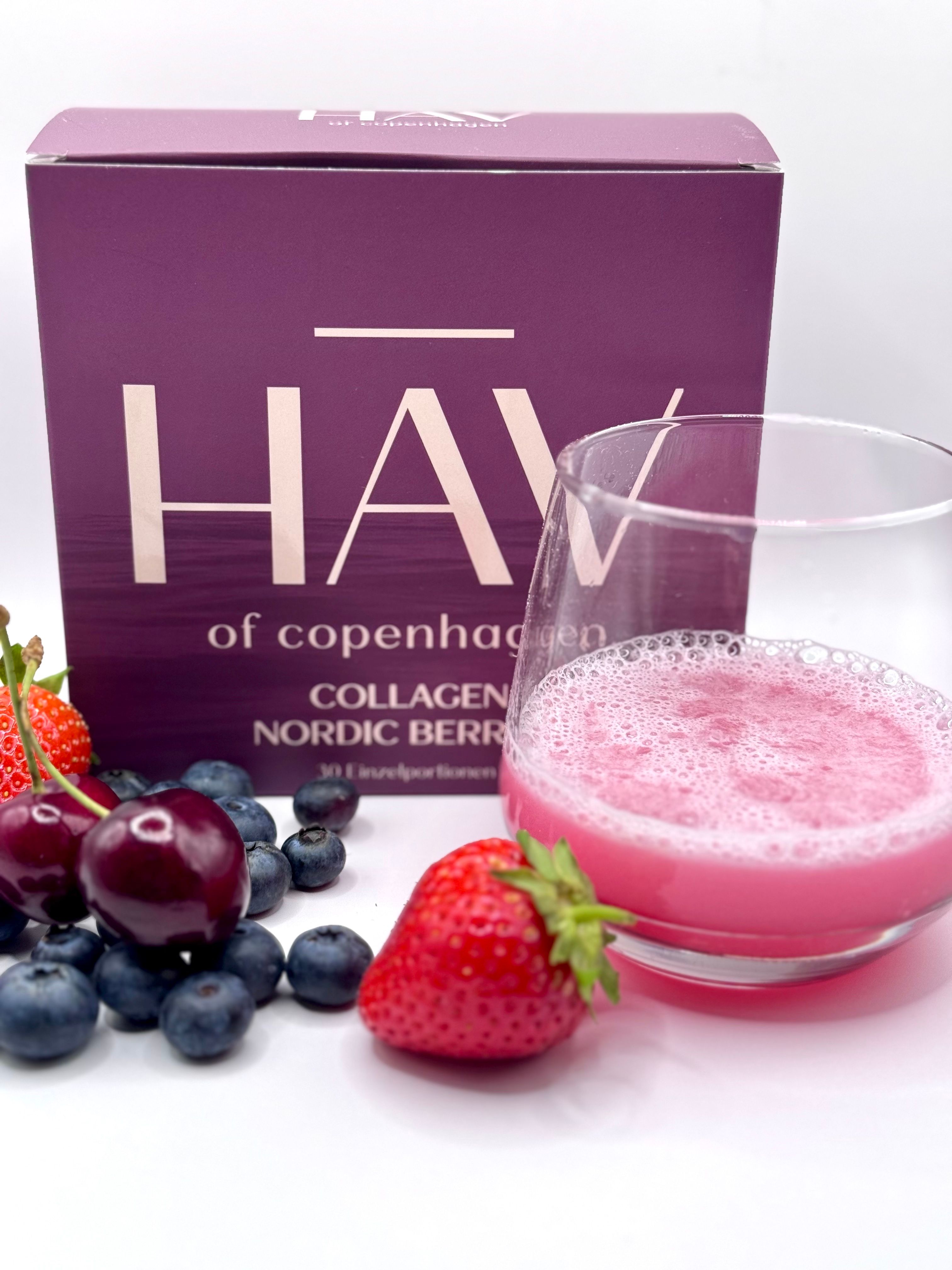 Produktverpackung und Glas mit rosa Flüssigkeit. Daneben Beeren: Erdbeeren, Blaubeeren, Kirschen.
