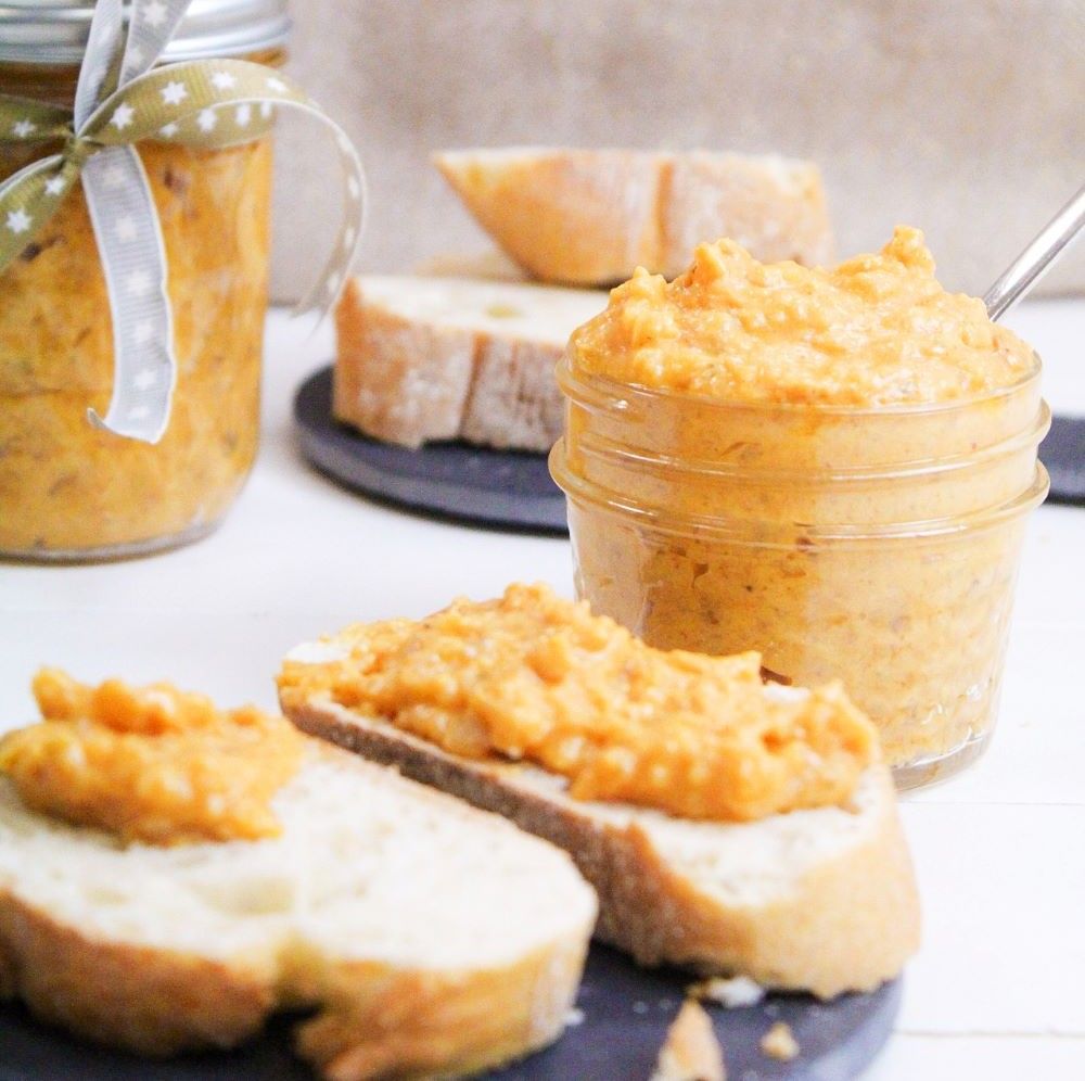 Gläser mit orangefarbener Paste, auf Brot serviert. Brotstücke und Löffel daneben. Ein Glas mit Schleife.