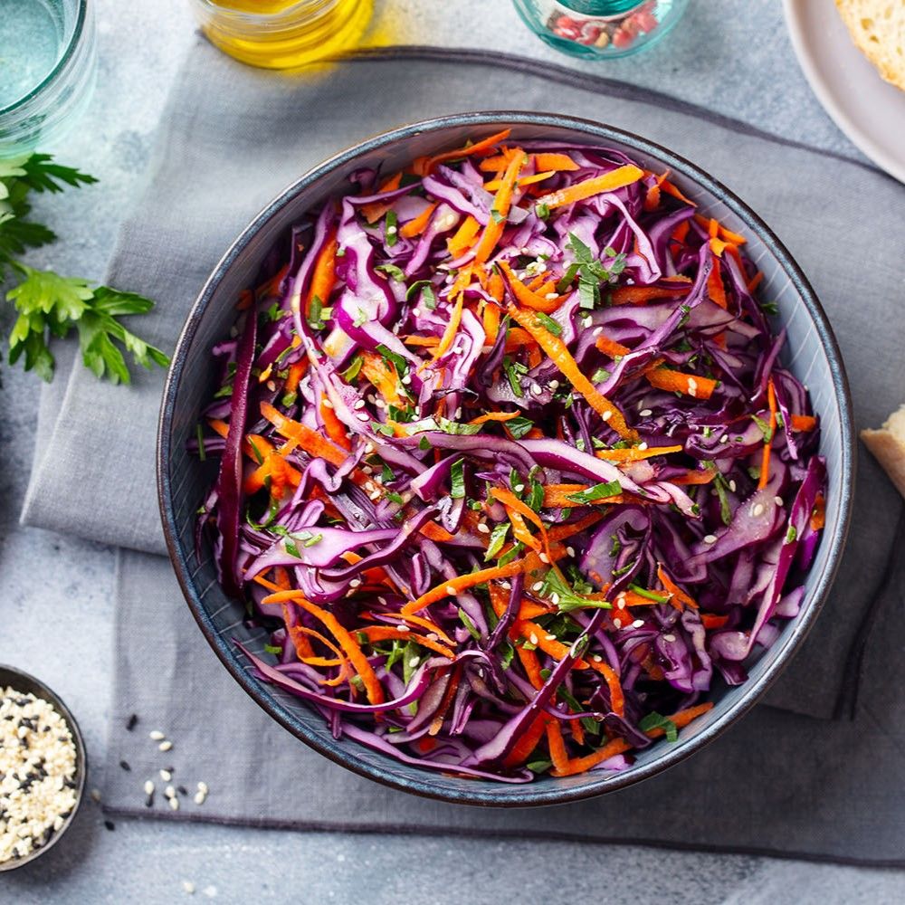 Nahaufnahme eines Salats mit Rotkohl, Karotten, Petersilie und Sesam in einer blauen Schüssel. Auf grauem Untergrund.