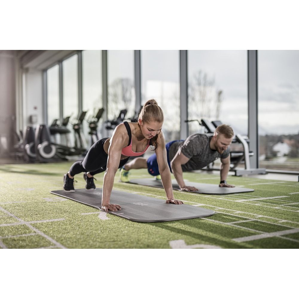 Zwei Personen trainieren auf grauen Fitnessmatten. Im Hintergrund Fitnessgeräte und Fenster.