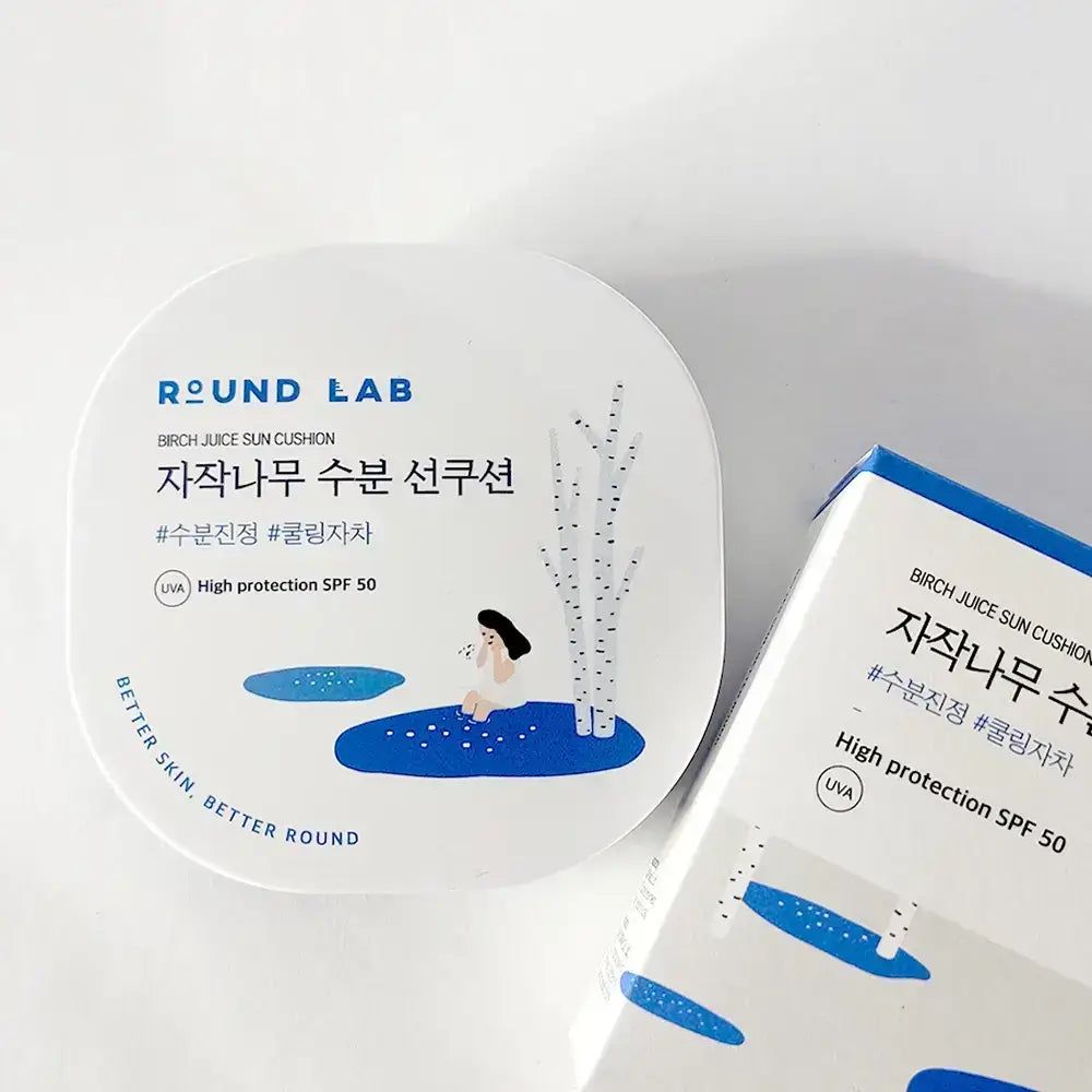 Weiße, quadratische Dose mit Text und Illustrationen. Aufschrift ROUND LAB, Birch Juice Sun Cushion. Daneben eine Schachtel.