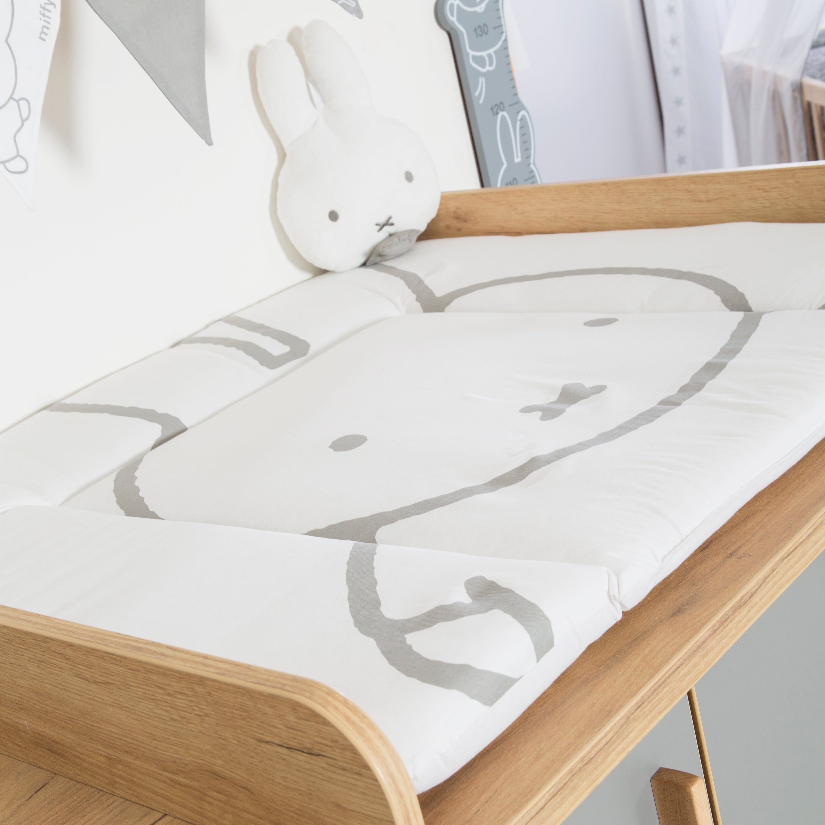 Wickelauflage auf Holzwickeltisch. Weiß mit grauem Miffy-Motiv. Stofftier und Dekoration im Hintergrund.