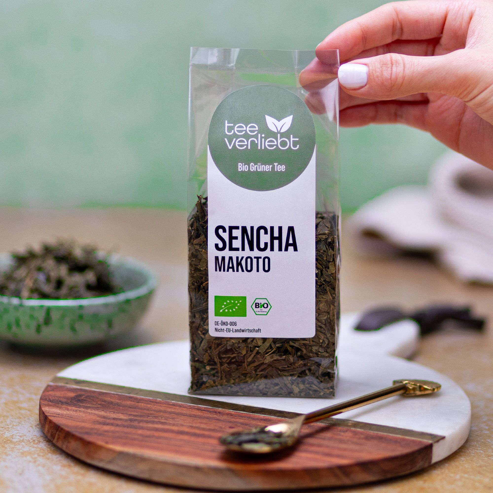 Hand hält eine Packung loser Grüntee. Auf einem Holzbrett mit Löffel. Aufdruck: teeverliebt, Sencha Makoto, Bio Grüner Tee.