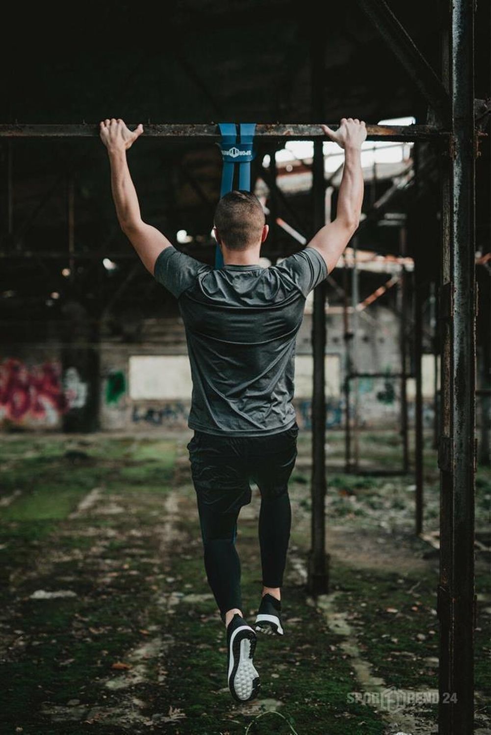 Mann trainiert an einer Stange mit blauem Fitnessband. Im Hintergrund eine Ruine.