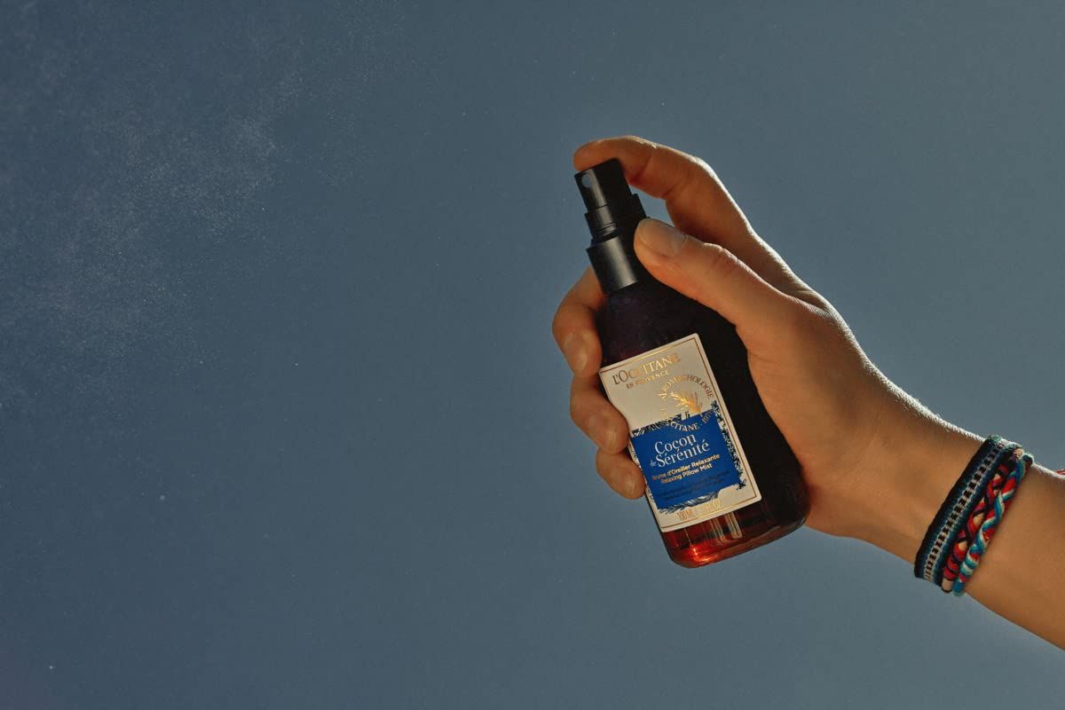 Hand hält braune Flasche mit schwarzem Sprühkopf. Sprühnebel sichtbar. Etikett mit Produktnamen. L'Occitane Aromachologie Kopfkissen-Nebel Entspannend.