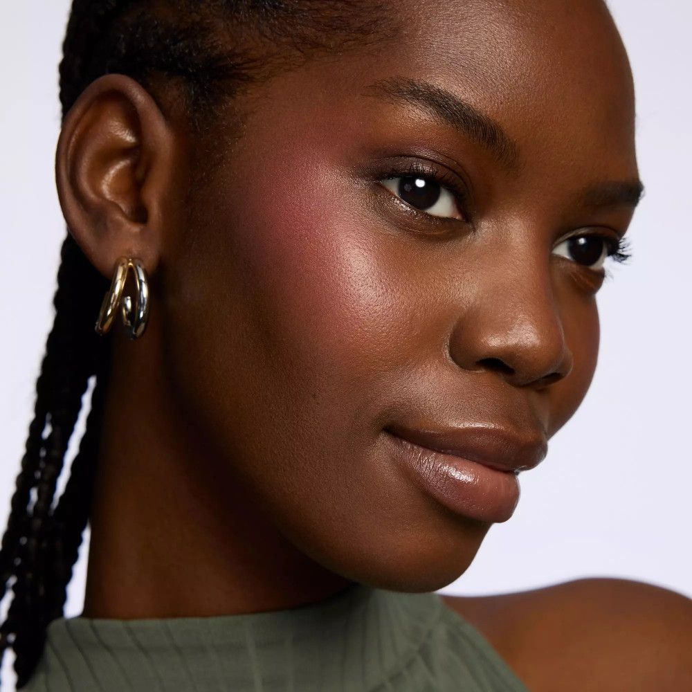 Porträt einer Frau mit Rouge auf den Wangen. Sie trägt Ohrringe. Dunkle Haut, schwarze Haare. Heller Hintergrund.