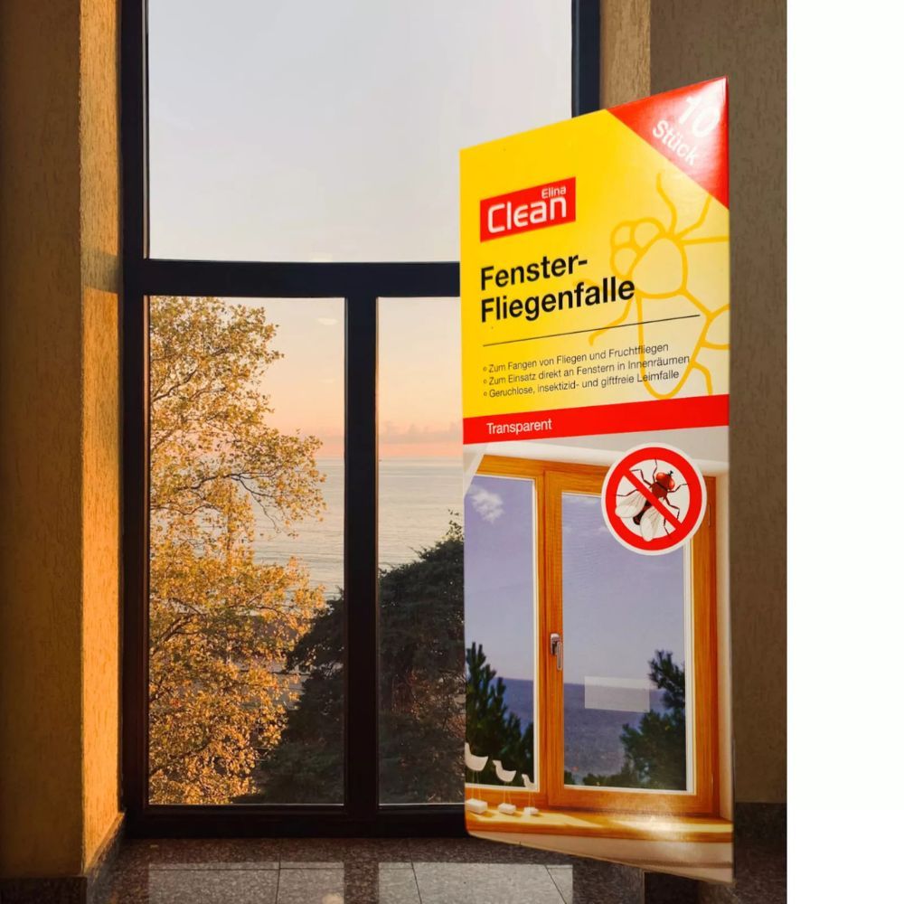 Elina Clean Fenster-Fliegenfalle Packung, gelb-rot, vor Fenster platziert. Produktname und Informationen sichtbar. 10 Stück. Fenster mit Ausblick.