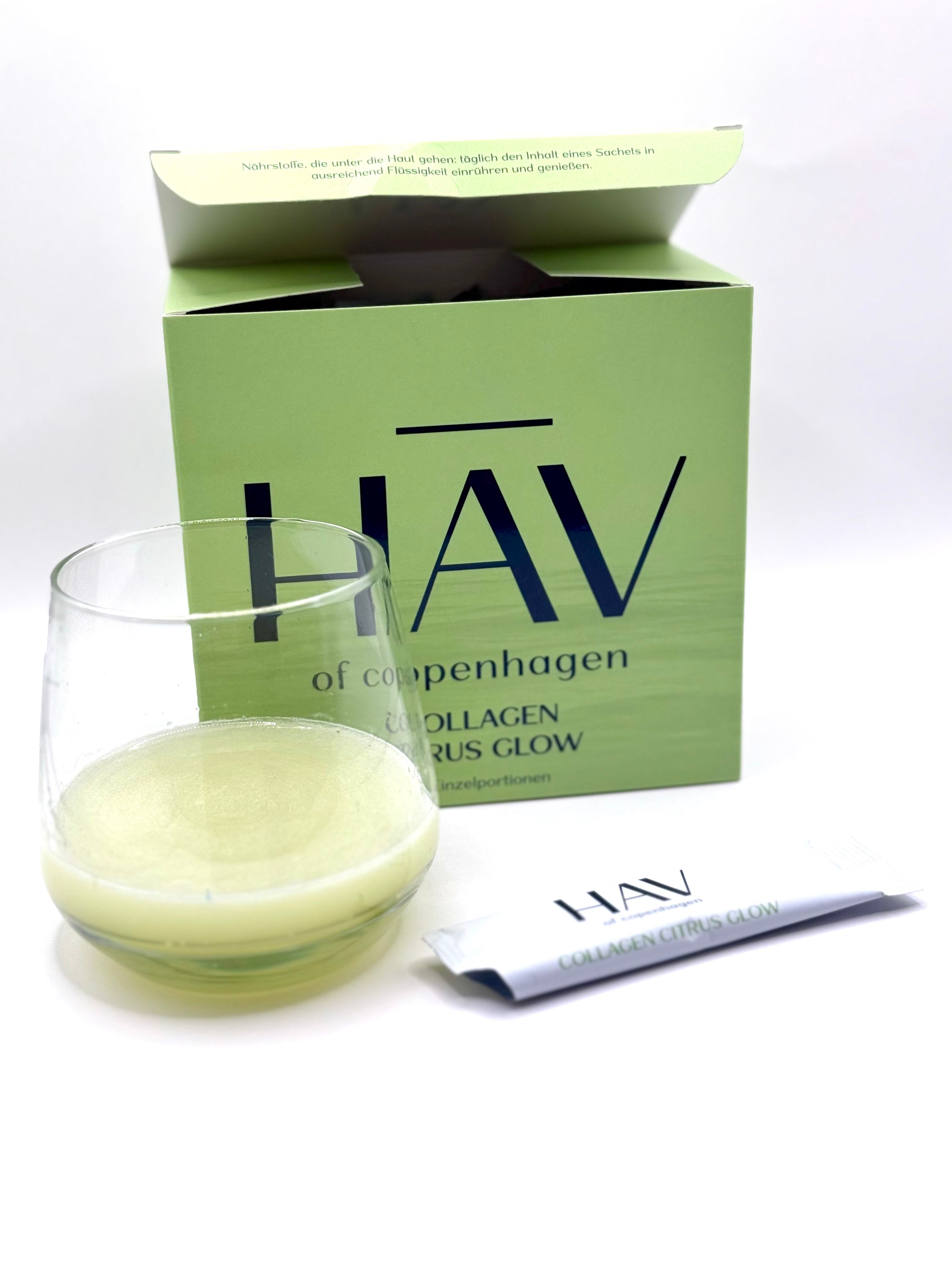 Glas mit Getränk und Einzelportion HAV Collagen Citrus Glow. Grüne Schachtel im Hintergrund.