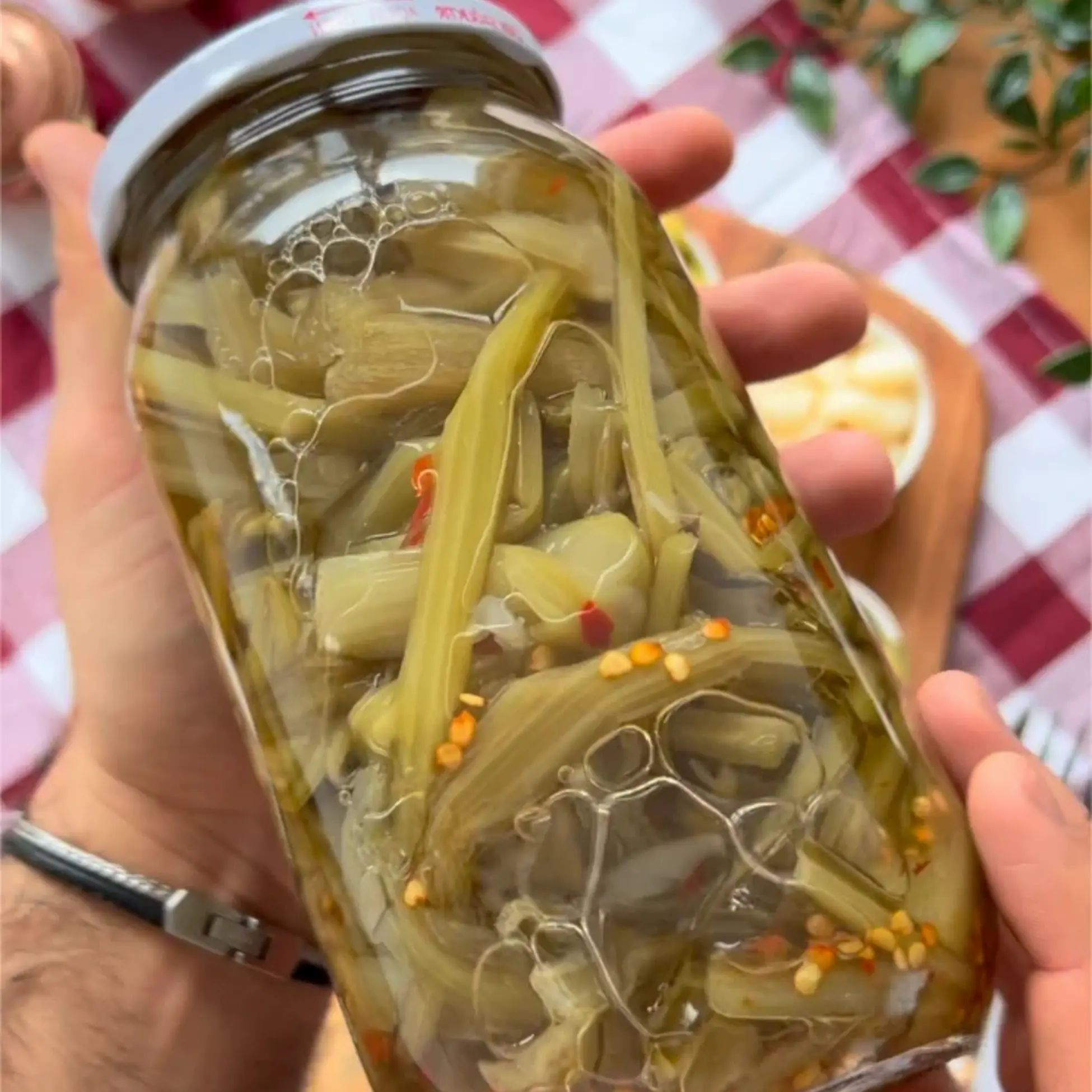 Glas mit eingelegten Pezik-Stangen, gehalten von Händen. Sichtbar sind Blasen im Glas. Hintergrund: karierte Tischdecke.