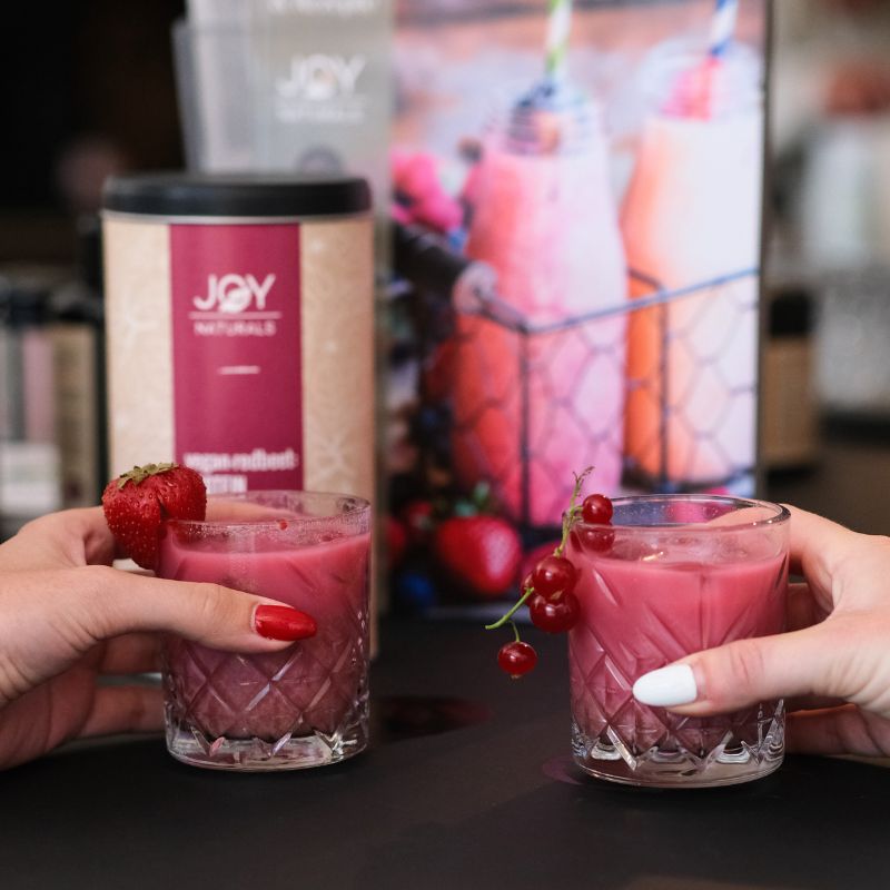 Zwei Gläser mit pinkfarbenem Getränk, gehalten von Händen. Erdbeere und Johannisbeeren als Garnitur. Dose im Hintergrund.