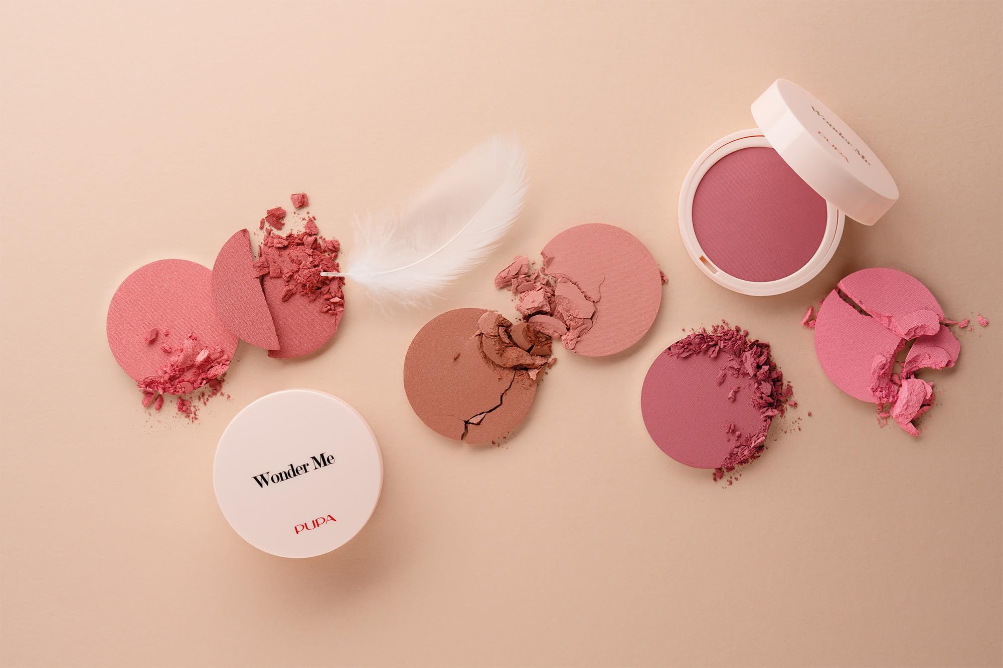 Mehrere runde Blush-Kompaktpuder, einige zerbrochen. Feder und Puder auf beigem Hintergrund. "Wonder Me" auf einem Deckel.