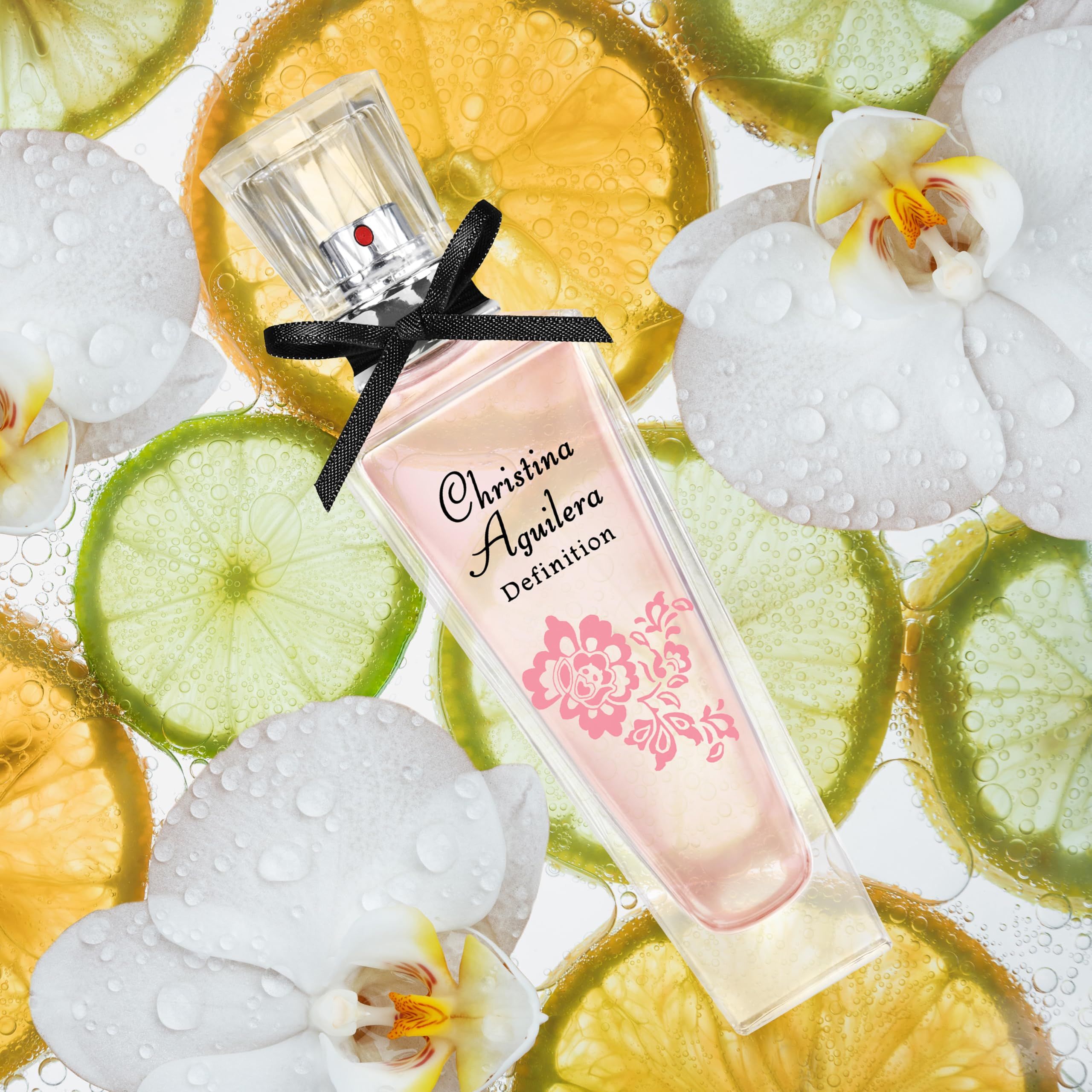Parfümflakon mit rosa Flüssigkeit, schwarzer Schleife und floralem Design. Aufschrift: Christina Aguilera Definition. Hintergrund: Früchte.