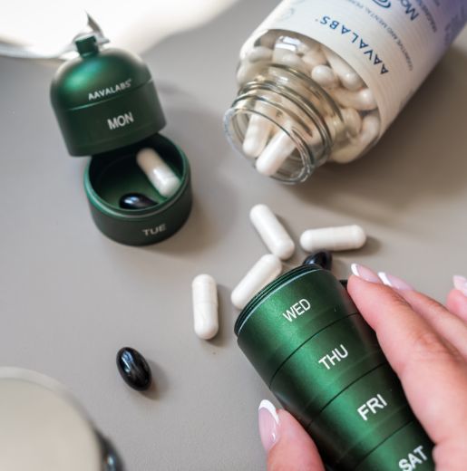Grüne Pillendose, ein Fach geöffnet mit weißen und schwarzen Pillen, daneben eine Pillenflasche.