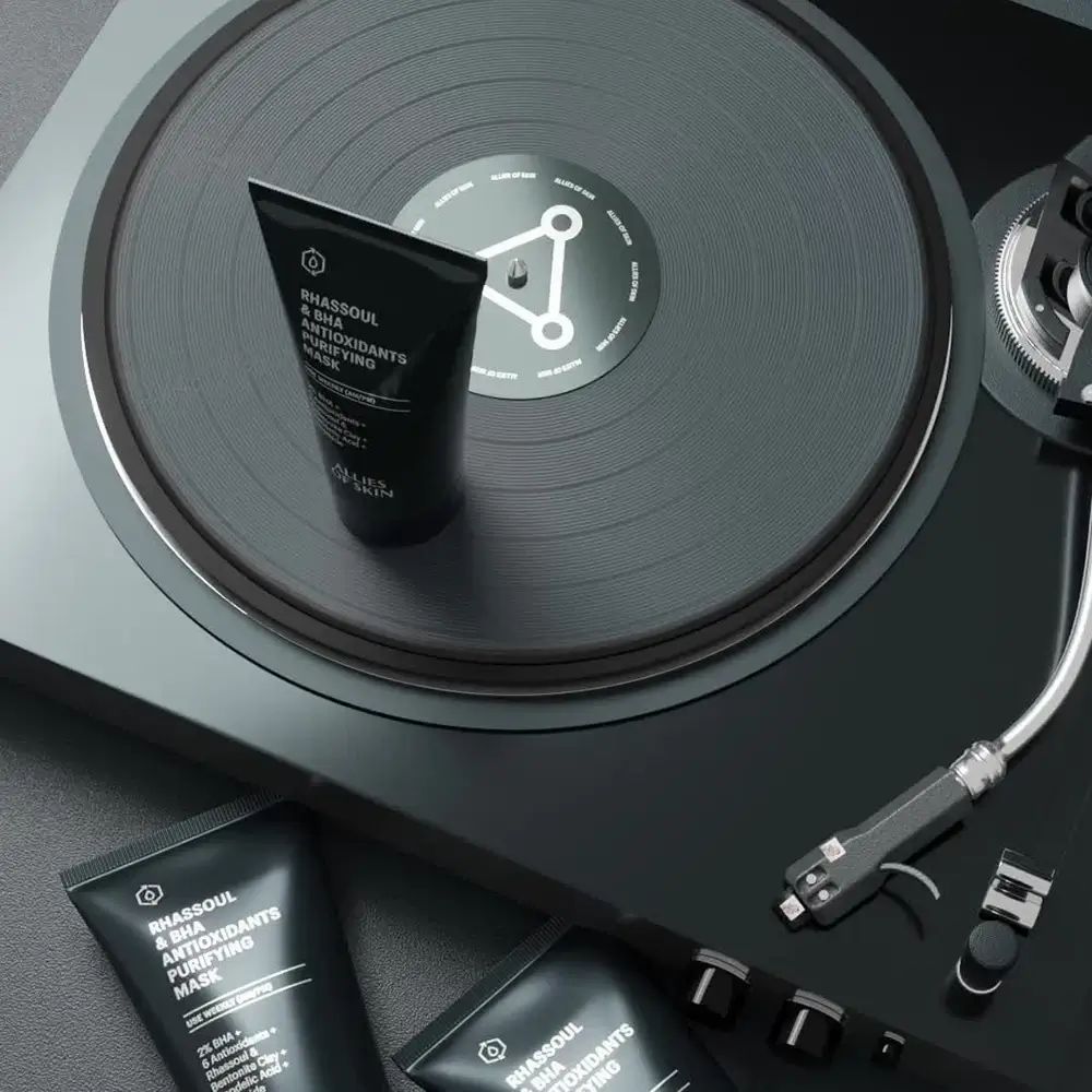 Drei schwarze Tuben auf einem Plattenspieler. Produktname und Marke sichtbar. Text: Rhassoul & BHA Antioxidants Purifying Mask. Marke: ALLIES OF SKIN.