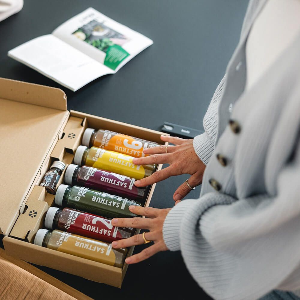 Geöffnete Verpackung mit 6 Flaschen Kloster Kitchen Saftkur. Eine Person berührt die Flaschen. Buch im Hintergrund.