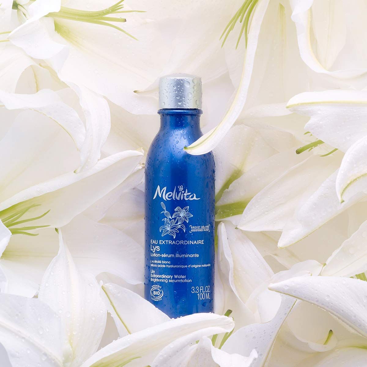 Blaue Flasche mit silbernem Deckel, umgeben von weißen Lilienblüten. Text: Melvita, Eau Extraordinaire Lys.