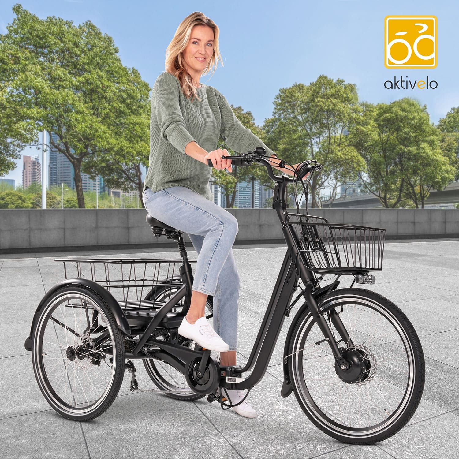 Frau fährt auf einem schwarzen Elektro-Dreirad. Logo 'aktivelo' sichtbar. Korb vorne. Hintergrund: Stadtlandschaft.