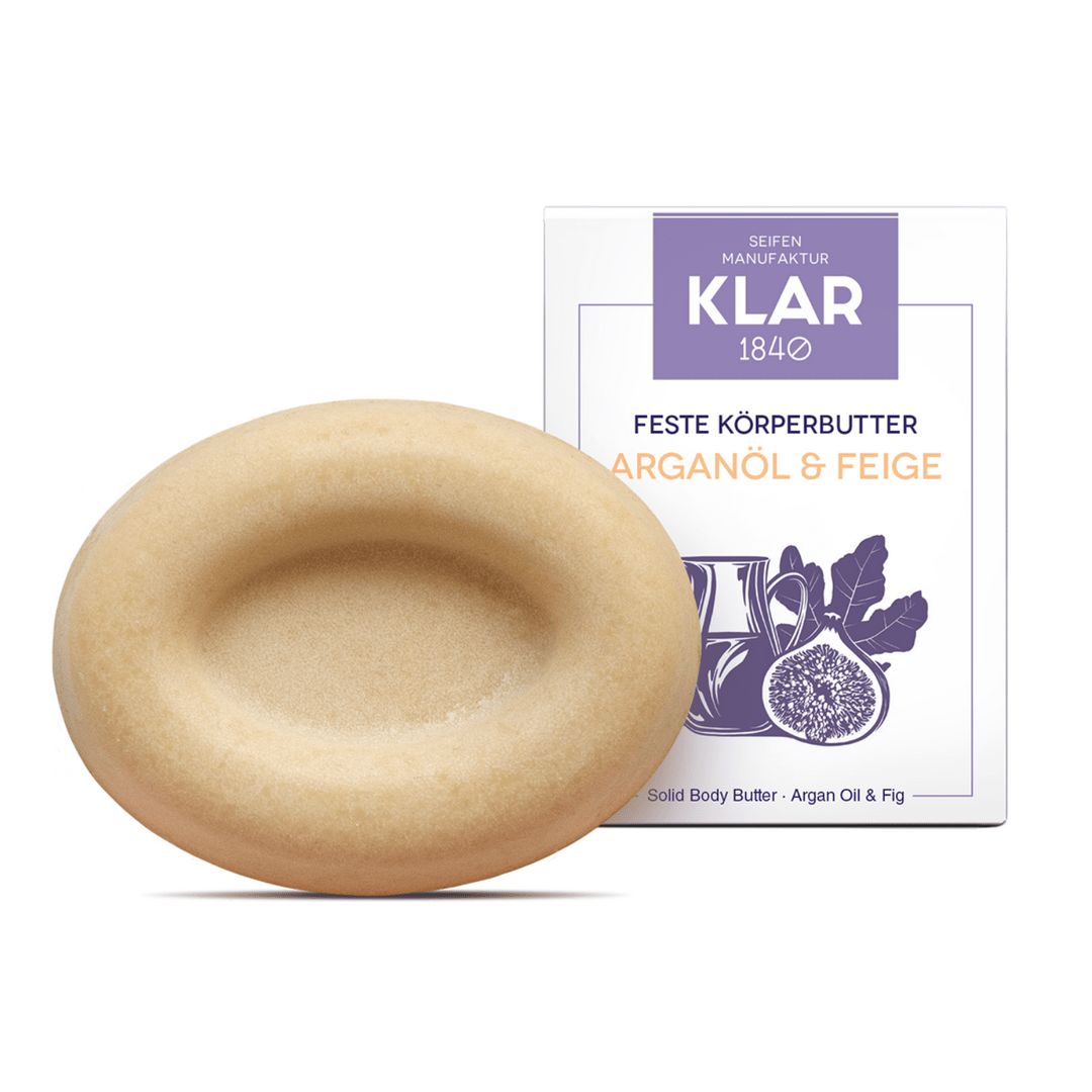Ovale, beige Körperbutter neben Verpackung. Die Butter hat eine konkave Form. Verpackung mit lila Logo und Produktbezeichnung: Feste Körperbutter Arganöl & Feige.