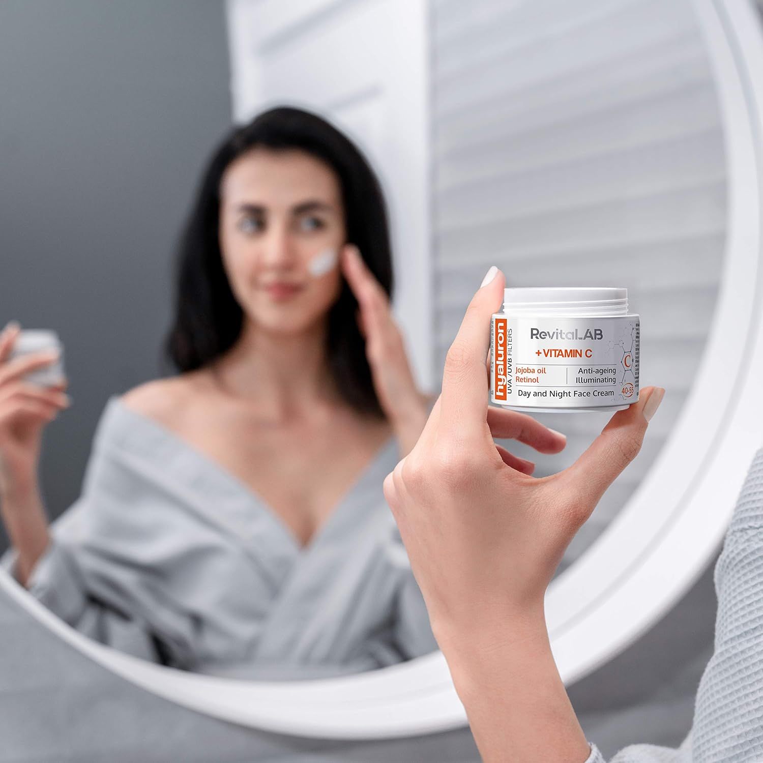 Frau im Spiegel, hält Tiegel RevitaLAB + Vitamin C. Aufschrift: Jojobaöl, Retinol, Anti-Aging, Tages- und Nachtcreme. Bademantel.