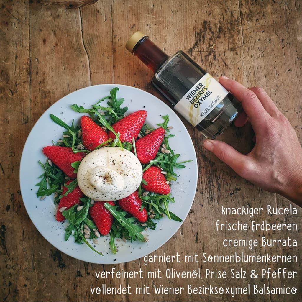 Eine Flasche Oxymel Balsamico. Glasflasche mit goldfarbenem Verschluss und Etikett. Daneben ein Teller mit Salat und Erdbeeren.