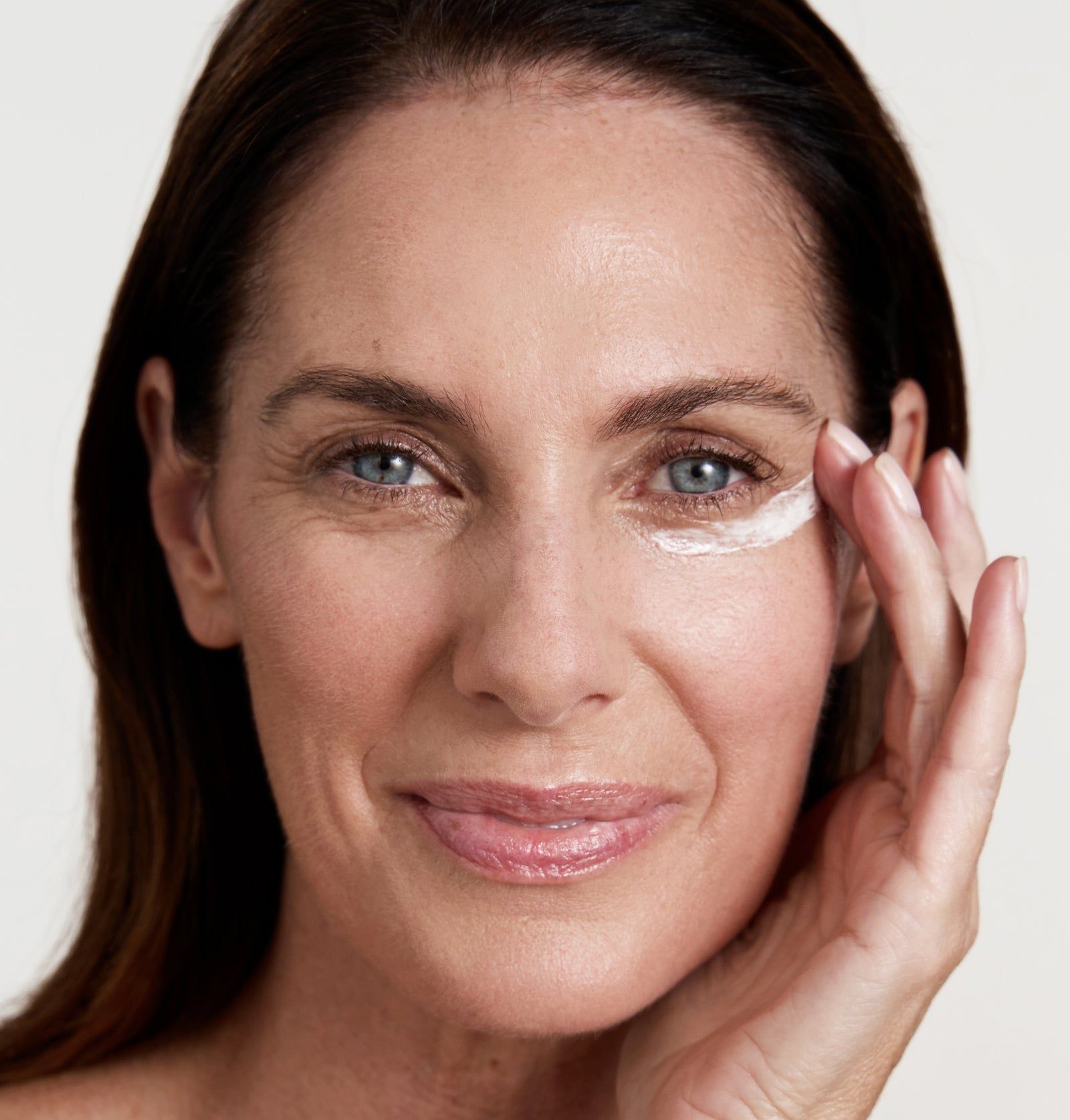 Frau mit Creme unter dem Auge. Die Hand berührt die Haut. Helle Haut, braune Haare, blauen Augen.