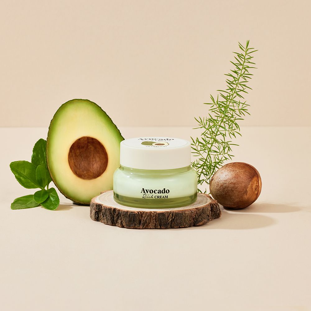 Cremebehälter auf Holzscheibe, neben Avocadohälfte, Blattwerk und Kern. Aufschrift: Avocado Rich CREAM.
