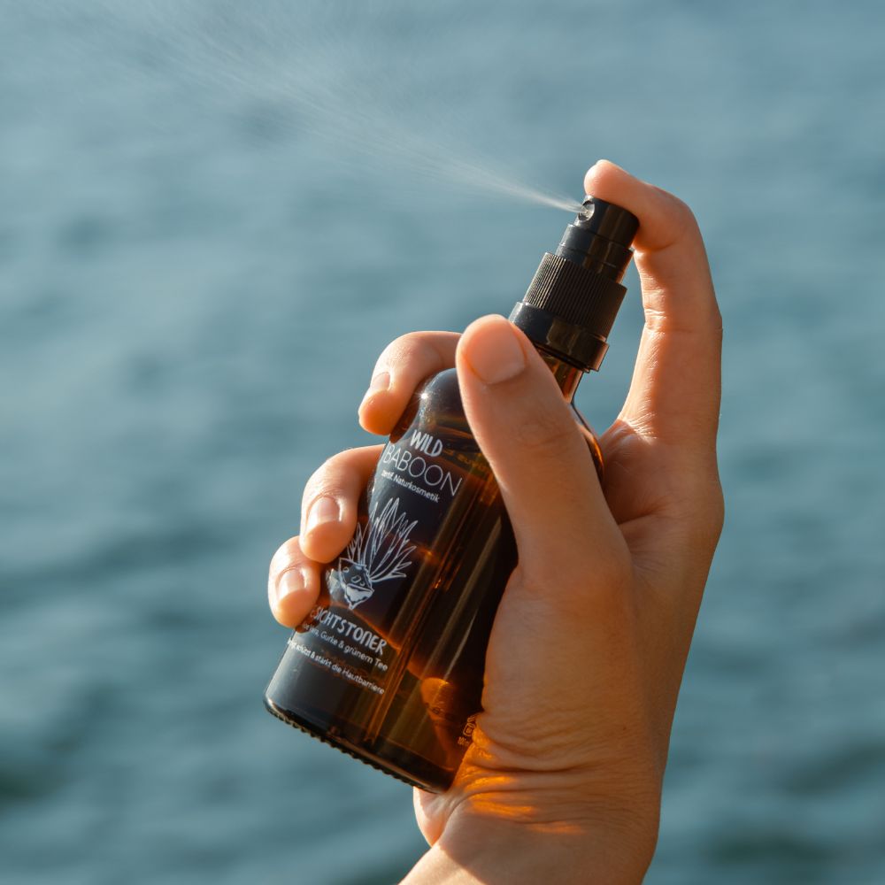 Person sprüht Gesichtstoner. Braune Flasche mit schwarzem Sprühkopf. Text: Wild Baboon Gesichtstoner. Wasser im Hintergrund.