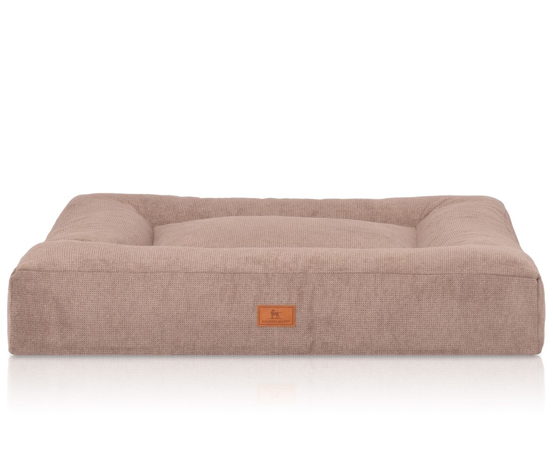 Rechteckiges Hundebett in Beige. Erhöhte Seiten, flache Liegefläche. Kleines Logo-Etikett an der Vorderseite. Stoffstruktur sichtbar.