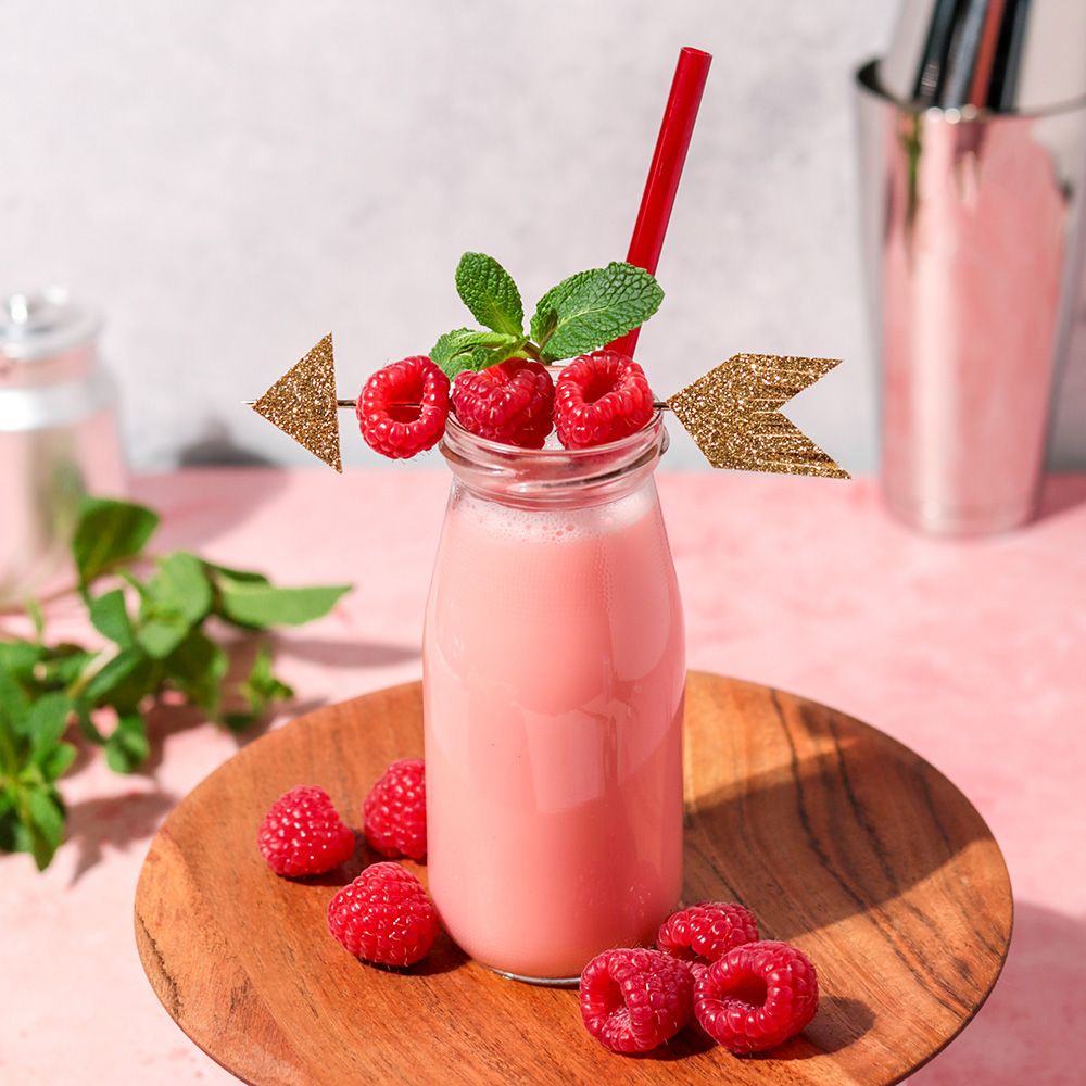 Glasflasche mit rosa Getränk, Himbeeren, Minze und Strohhalm. Auf Holztablett. Dekoration: goldener Pfeil.
