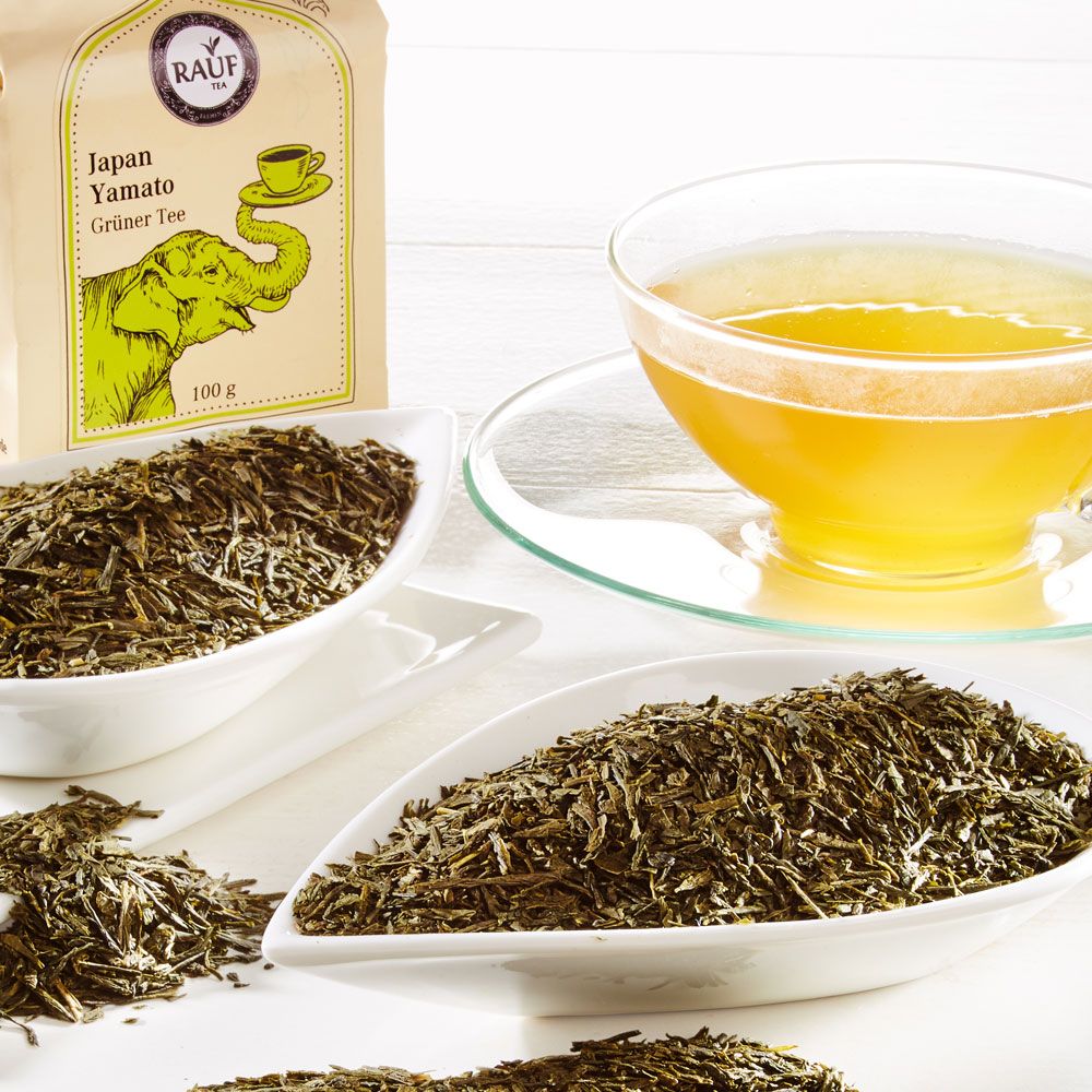 Schalen mit losem grünem Tee neben einer Tasse Tee und der Verpackung von Rauf Tee Japan Yamato Grüner Tee.
