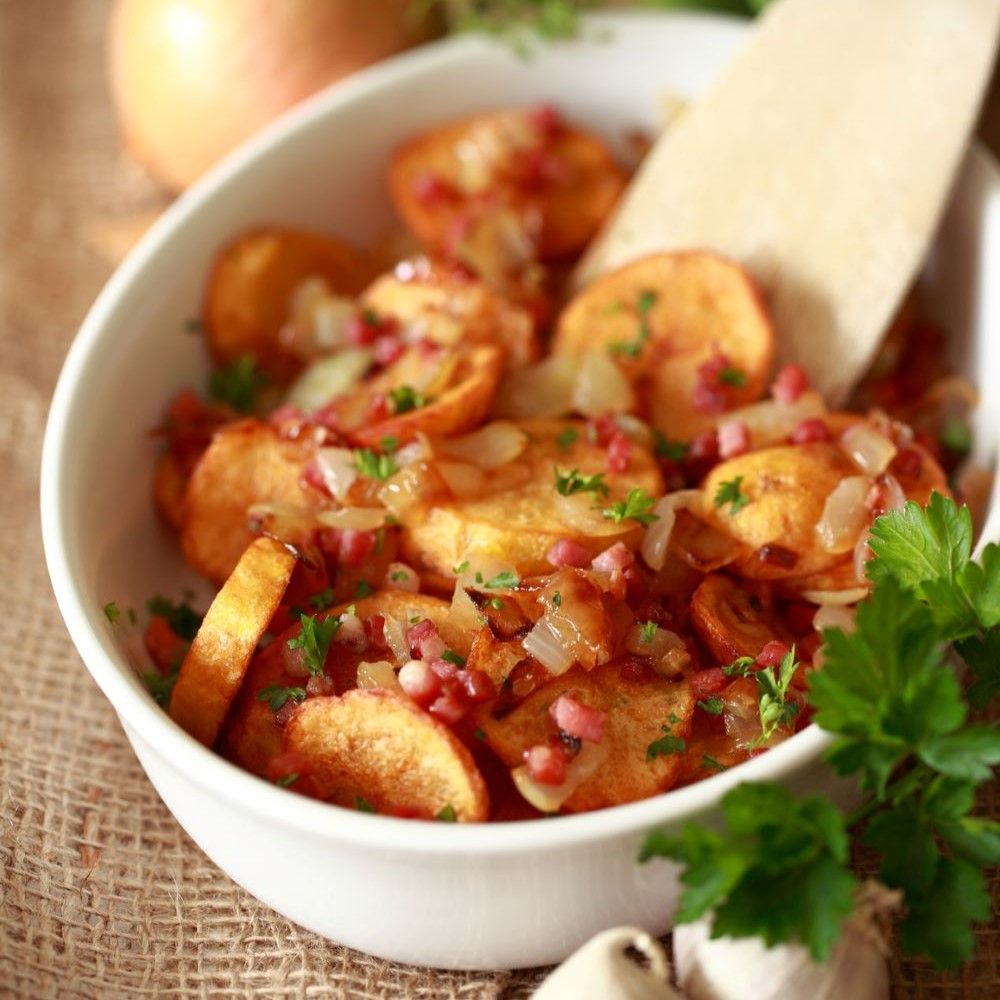 Gebratene Kartoffeln in einer weißen Schüssel. Mit Zwiebeln, Speck und Petersilie garniert. Auf einem groben Untergrund.