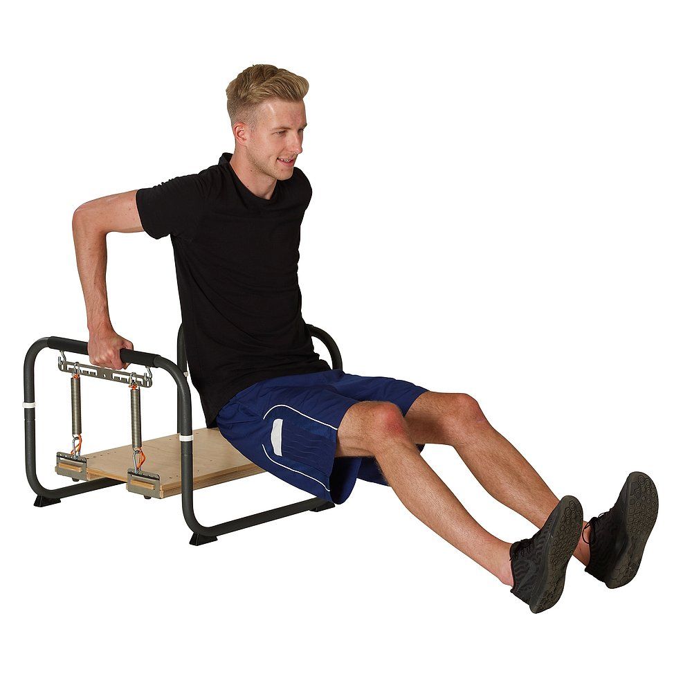 Mann sitzt auf Trainingsgerät. Schwarzer Rahmen, Holzplatte, Federn und Griffe. Mann in Sportkleidung. Weißer Hintergrund.