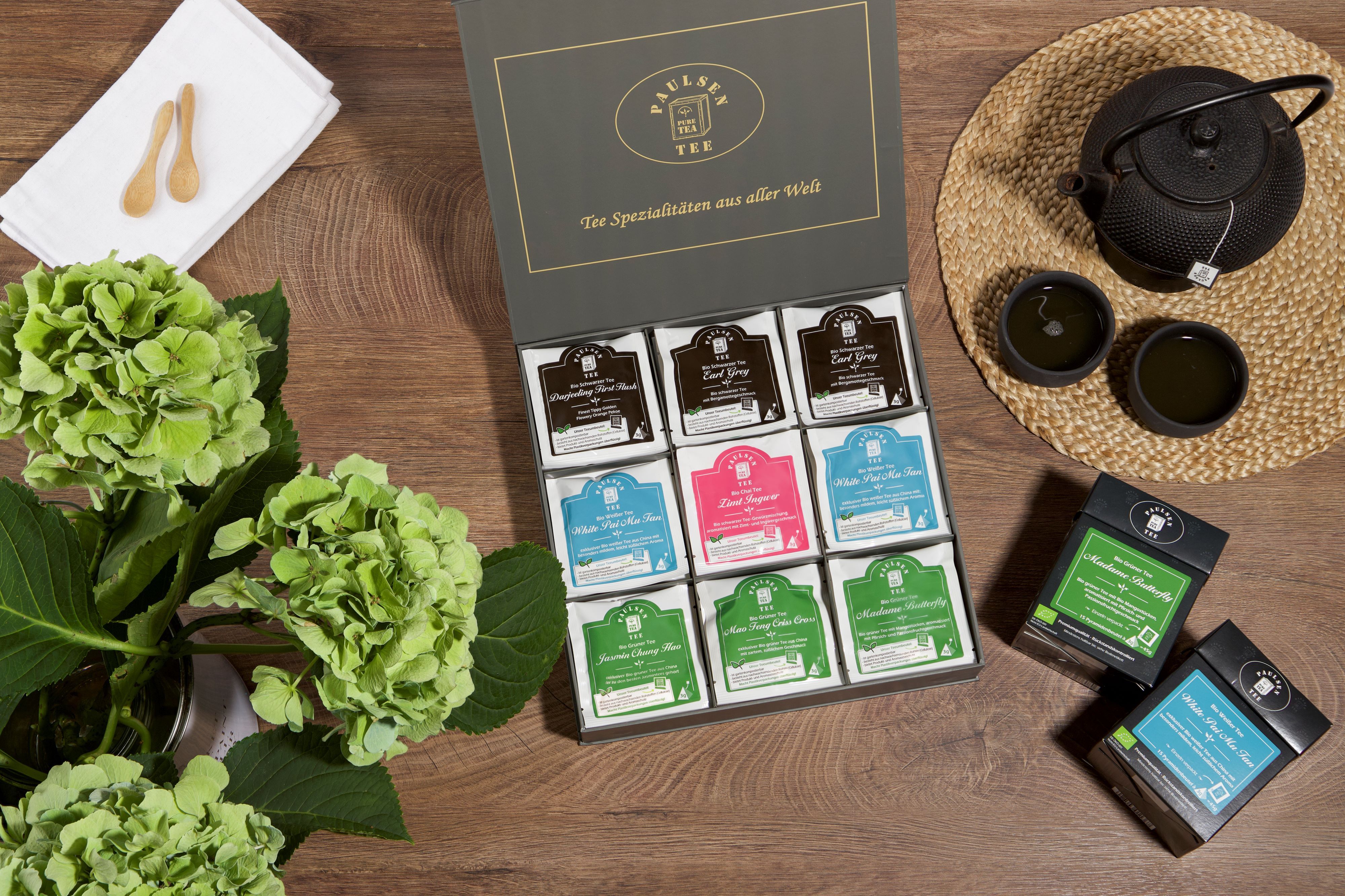 Geöffnete Geschenkbox mit Teebeuteln, daneben Tasse, Kanne, Blumen und weitere Teebeutel. Aufschrift auf der Box.