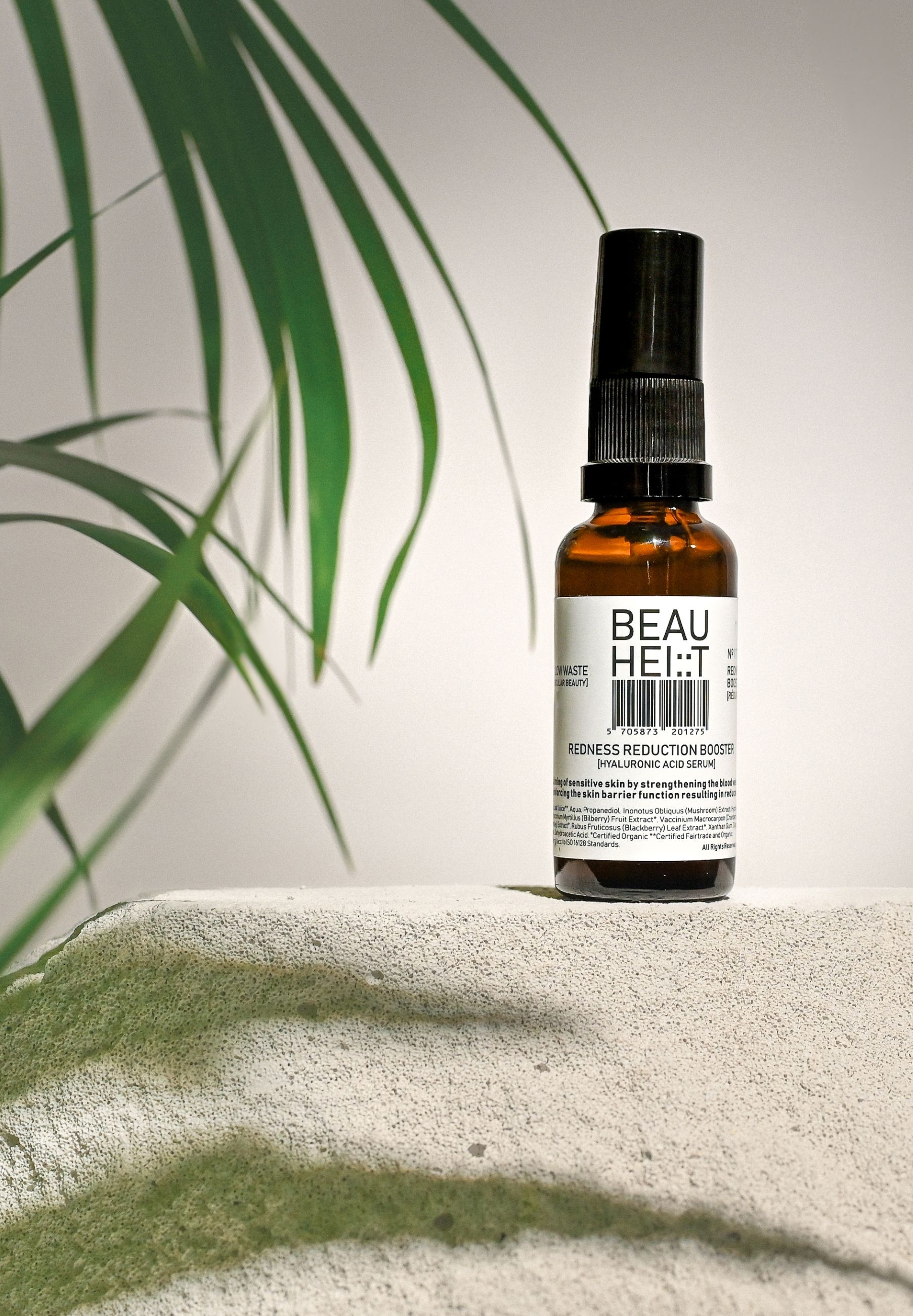 Braune Glasflasche mit schwarzem Sprühkopf auf einer sandfarbenen Oberfläche. Etikett mit Produktnamen. Grüne Pflanze im Hintergrund.