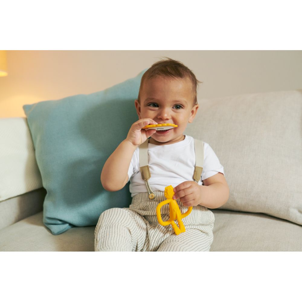 Baby sitzt auf Sofa und beißt auf gelben Beißring. Baby trägt Latzhose und weißes Shirt.