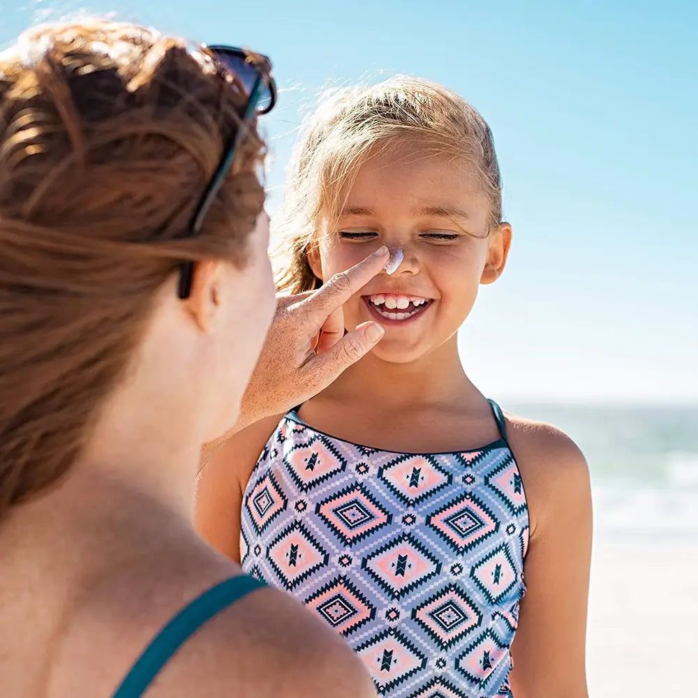 Mutter trägt Sonnenschutz auf die Nase ihres Kindes auf. Das Kind trägt einen Badeanzug am Strand.