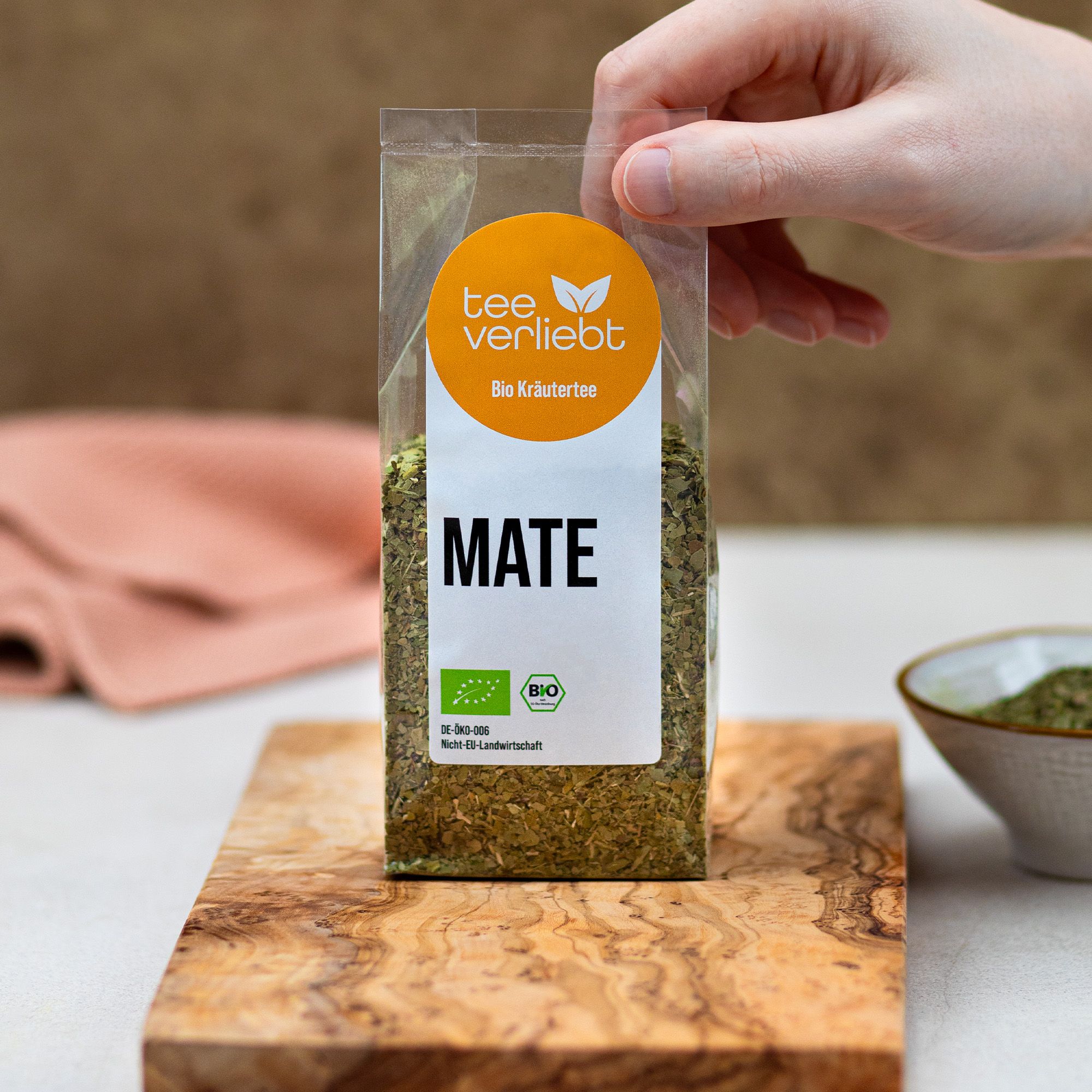 Hand hält transparente Verpackung mit losem Mate-Tee. Aufkleber mit Logo, Text 'Bio Kräutertee' und 'MATE'. Bio-Siegel.