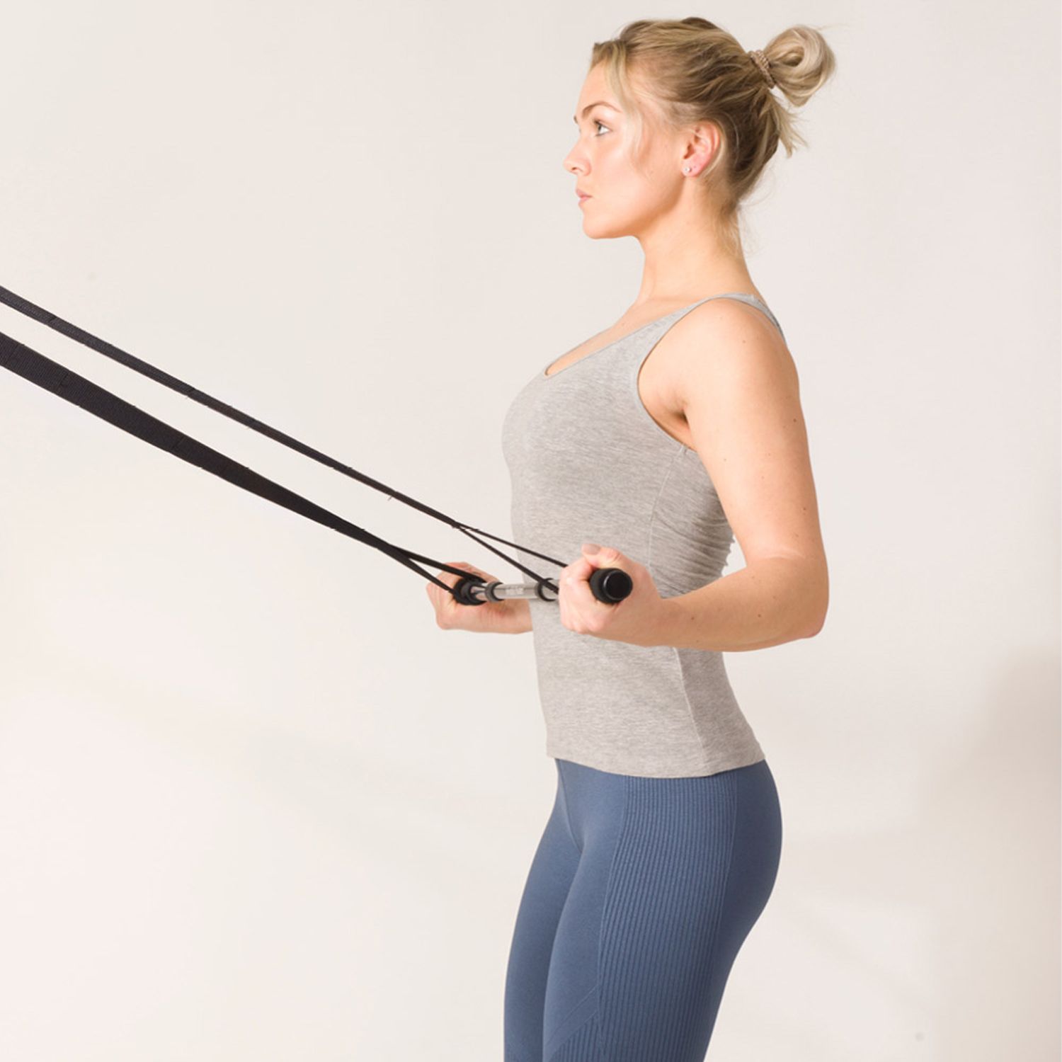 Frau mit Trainingsgerät. Schwarze Stange mit Bändern. Graues Oberteil, blaue Hose. Weißer Hintergrund.