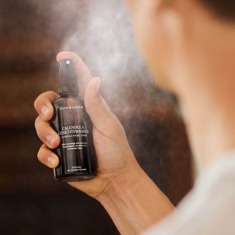 Hand hält JUNGLÜCK Calendula Gesichtswasser Spray und sprüht es auf das Gesicht.