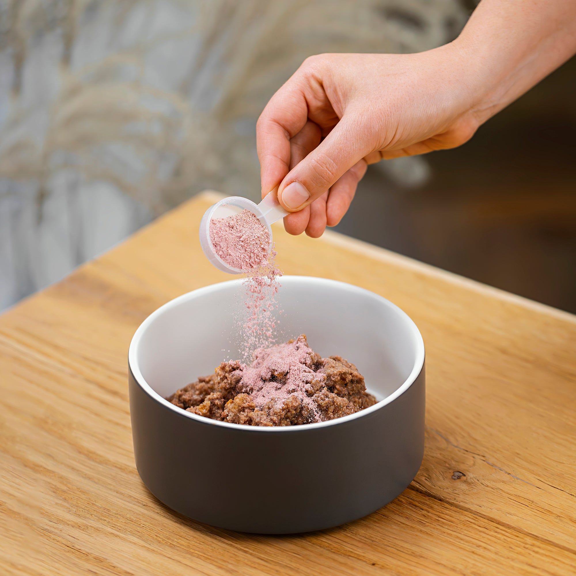 Hand schüttet rosa Pulver mit Messlöffel in eine Schüssel mit Futter. Holzoberfläche.