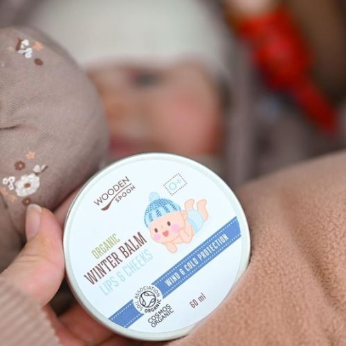 Runde Dose mit Deckel, gehalten von einer Hand. Aufschrift: Wooden Spoon, Organic Winter Balm Lips & Cheeks. Baby-Illustration. Eco Cert Cosmos Organic.