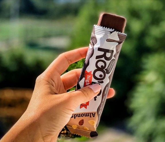 Hand hält teilweise geöffnete Riegelverpackung. Aufschrift: Roo'bar, high protein, Mandeln. Hintergrund: verschwommene Natur.