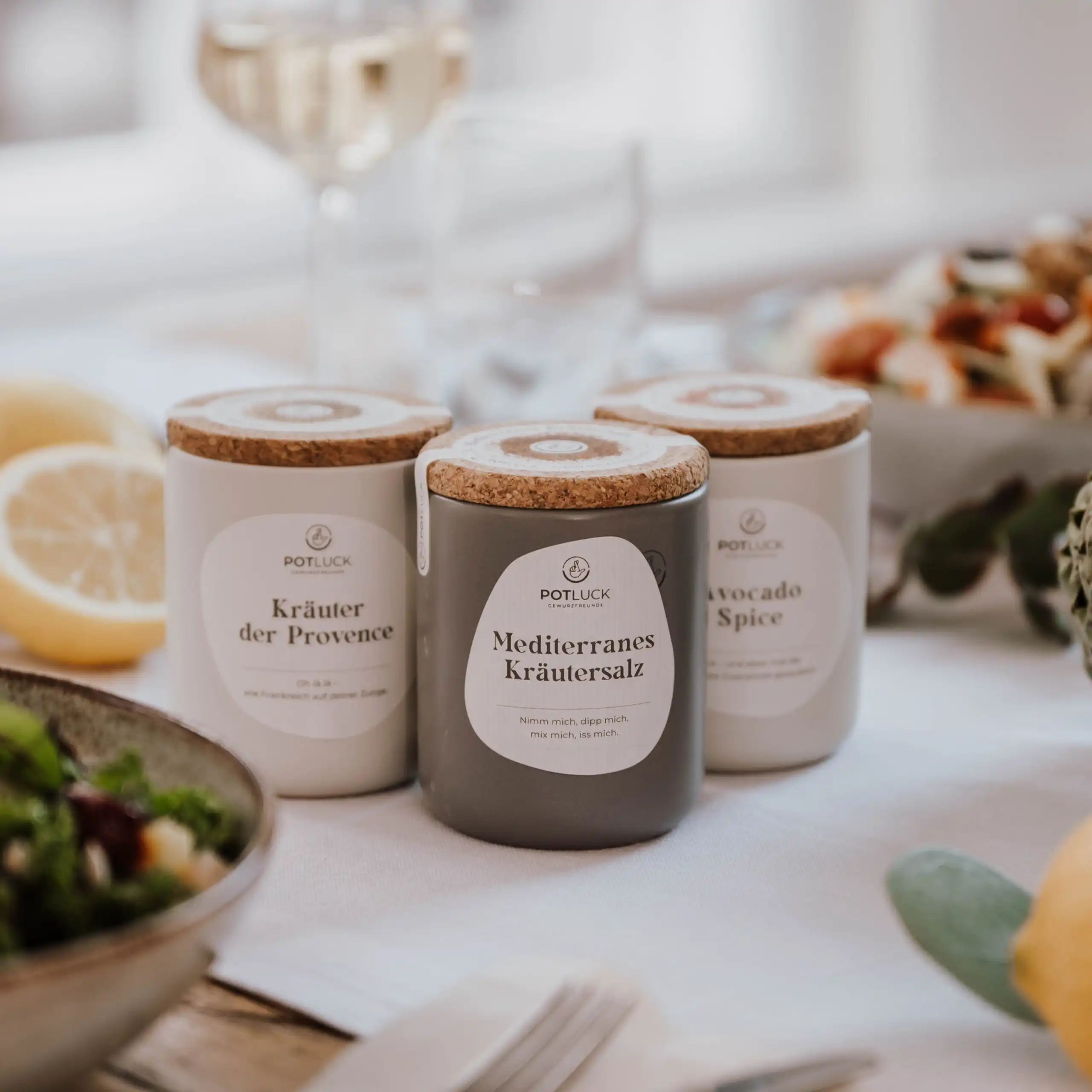 Drei Keramikbehälter mit Korkdeckeln. Etiketten mit "Kräuter der Provence", "Mediterranes Kräutersalz", "Avocado Spice".