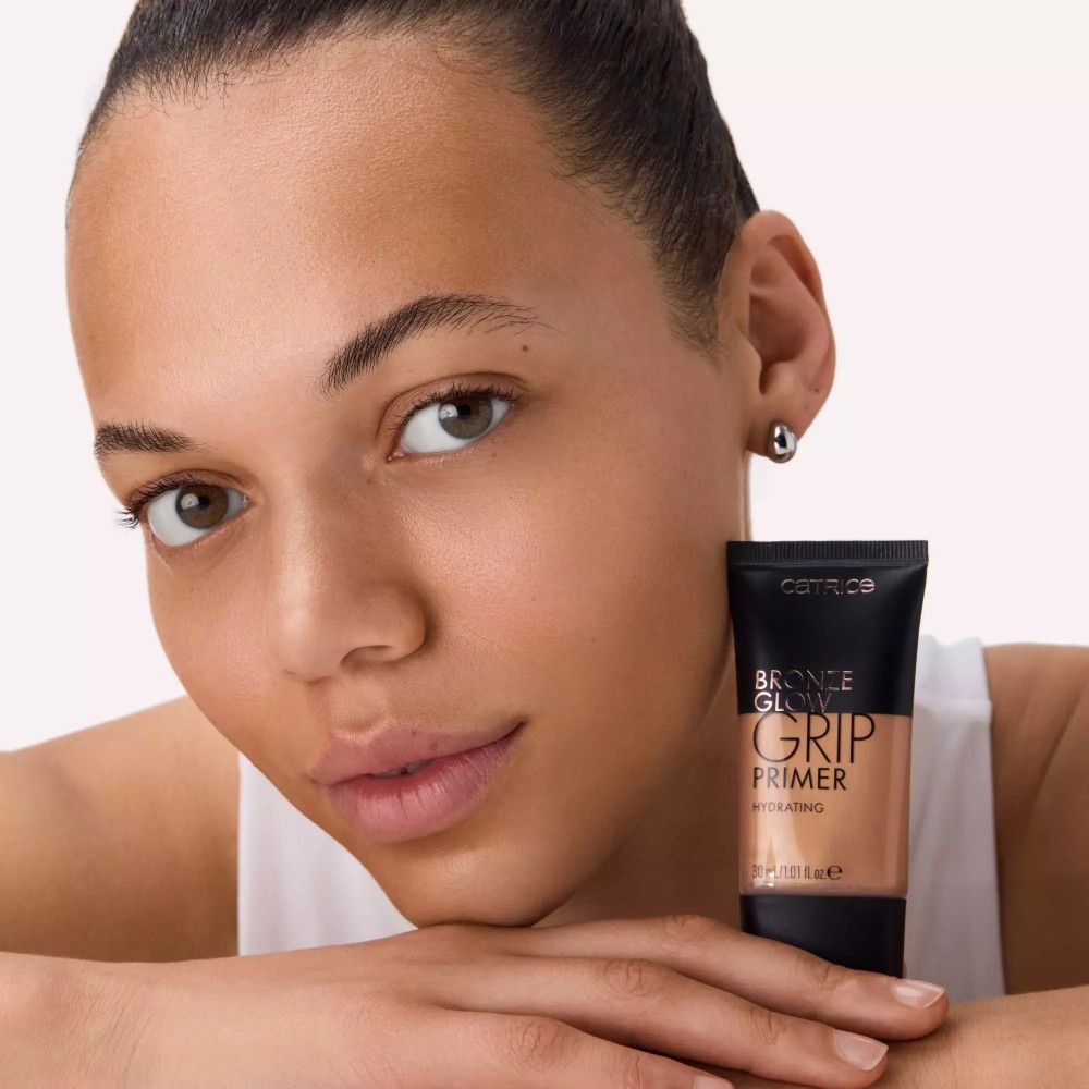 Frau hält Catrice Bronze Glow Grip Primer. Schwarze Tube mit bronzefarbener Flüssigkeit. Schriftzug: Catrice, Bronze Glow, Grip Primer, Hydrating.