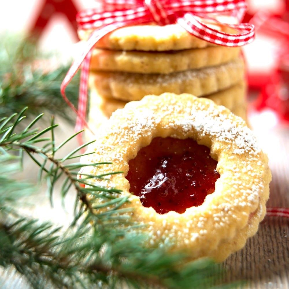 Weihnachtsplätzchen mit roter Füllung, Puderzucker und Tannenzweig. Stapel Kekse im Hintergrund, mit Schleife verziert.
